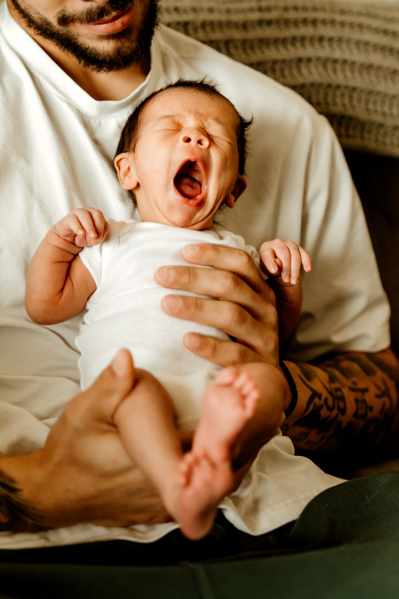 Newborn baby in a white onesie yawning widely while being held in the lap of a tattooed man in a white shirt.
