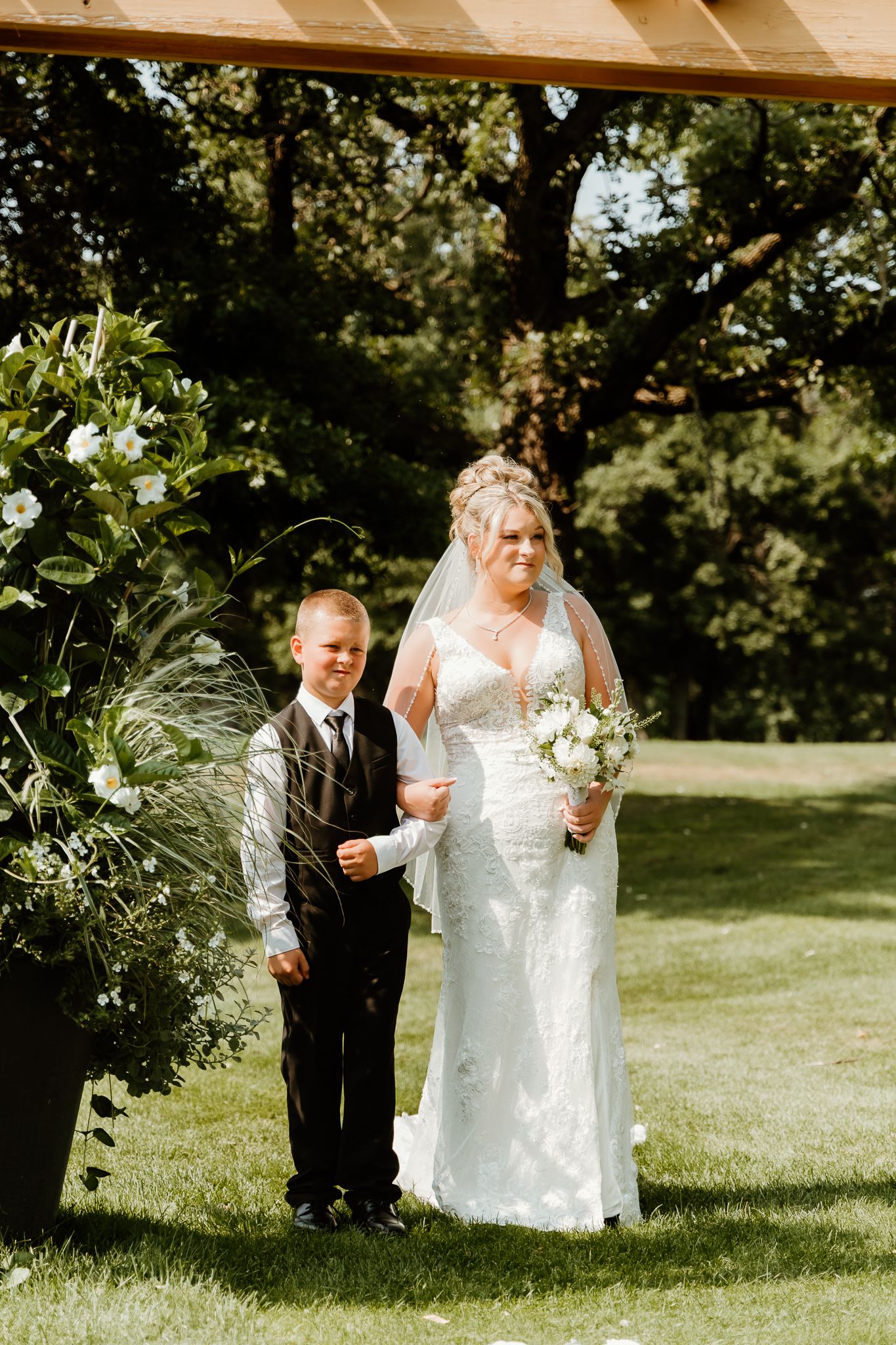 Bride in white lace gown holding bouquet walks with young boy in suit at outdoor wedding ceremony.