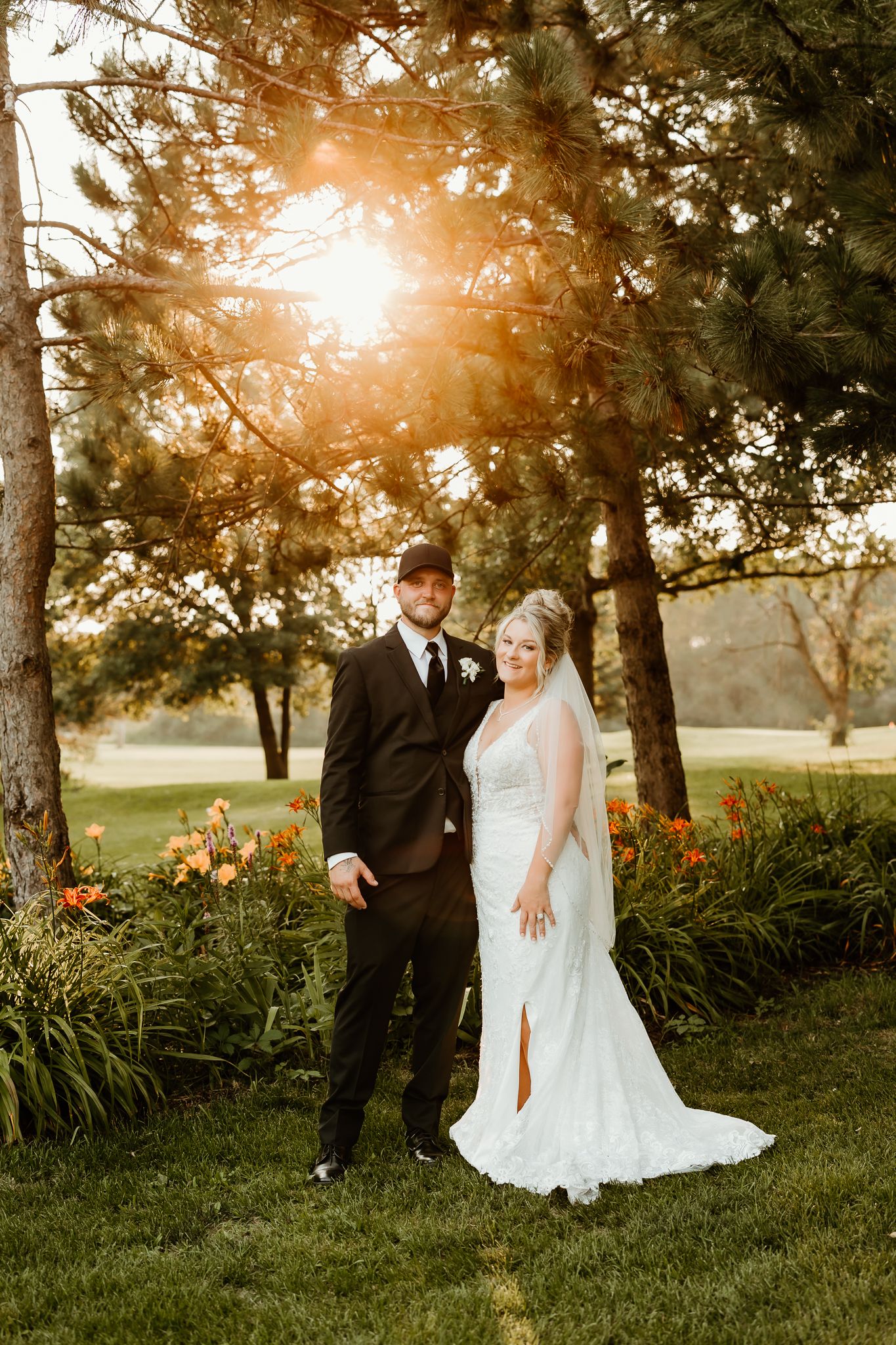Bride and groom pose together at golden hour under pine trees with orange flowers in a lush garden setting.