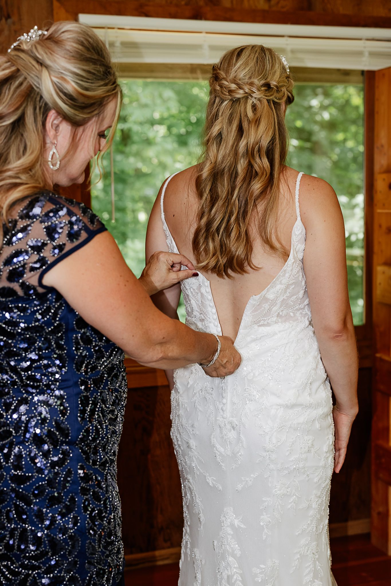 A woman in a navy sequin dress buttons the back of a bride's white lace wedding gown near a sunlit window.
