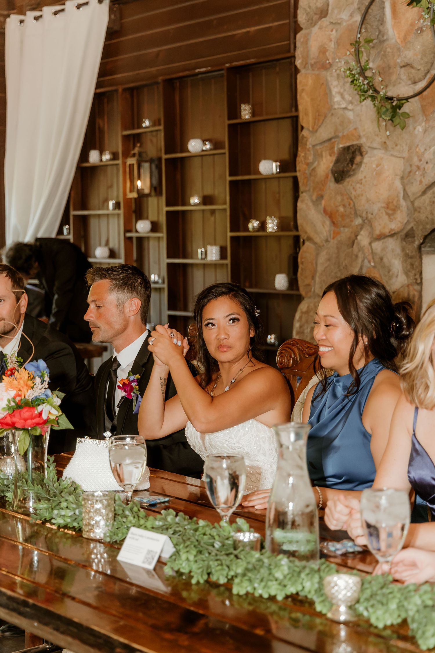 Bride and wedding party seated at decorated head table in rustic venue with stone fireplace and wooden shelving.
