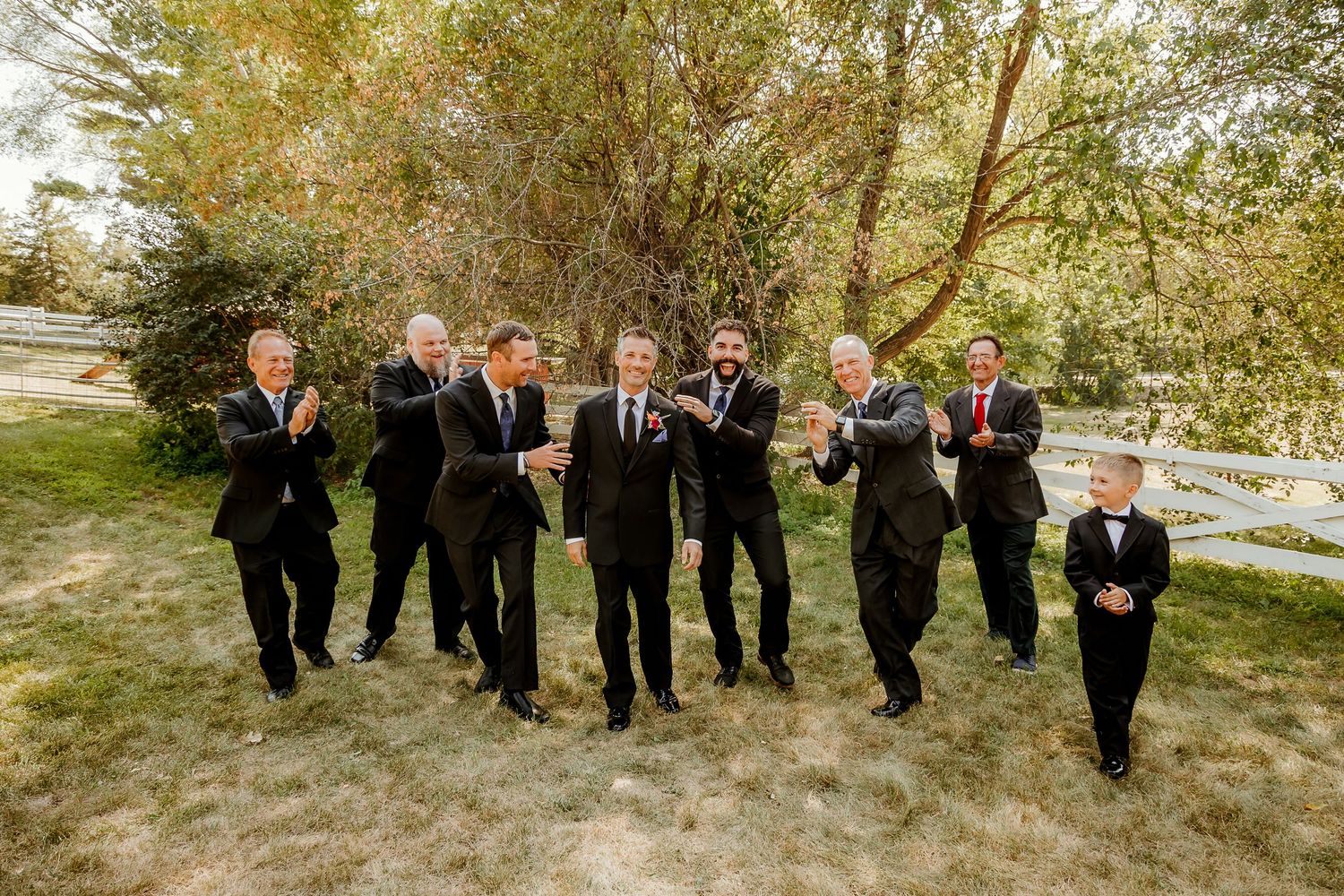 Groomsmen in black tuxedos toasting with champagne outdoors among trees at a wedding celebration.