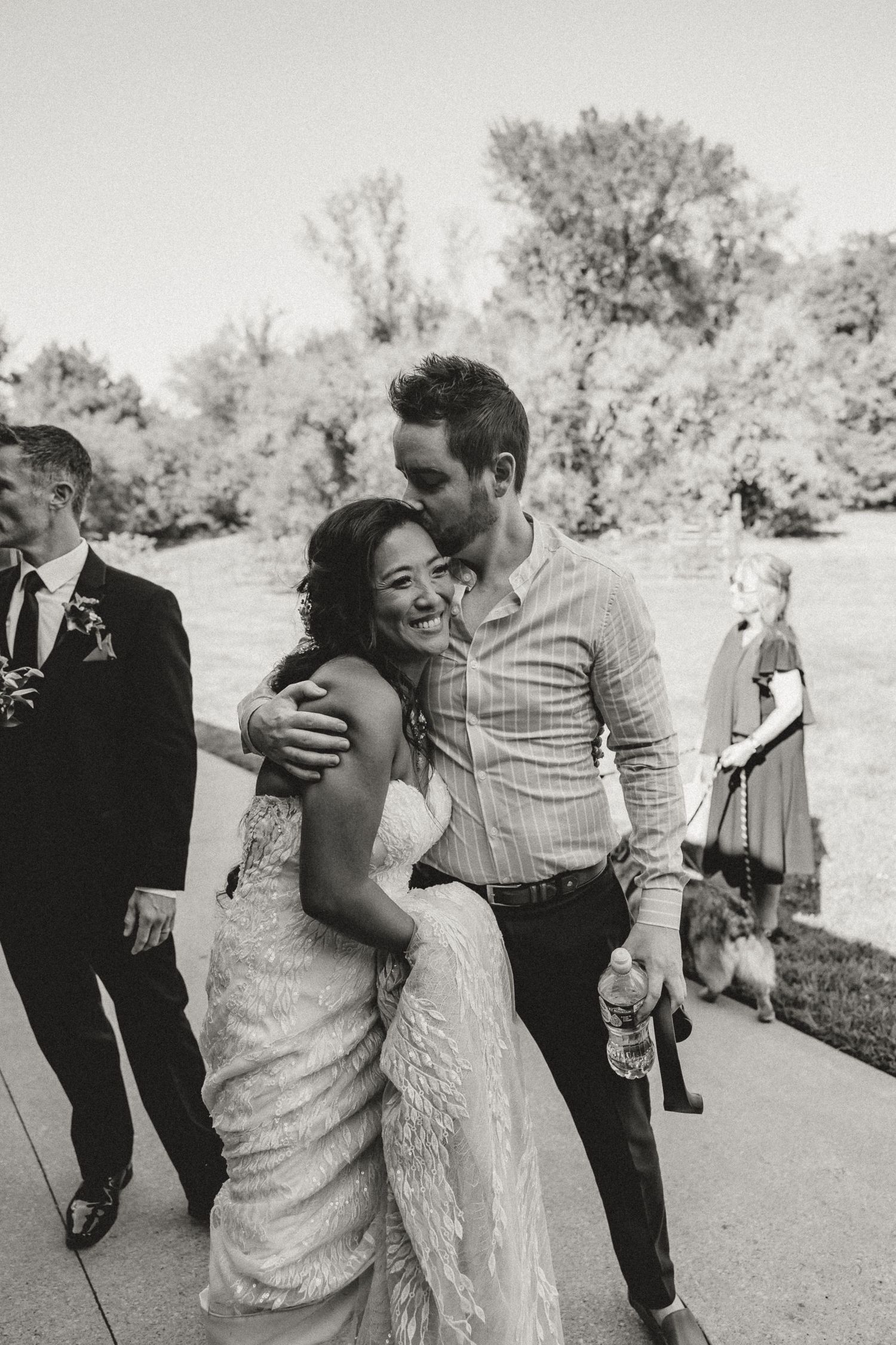Black and white photo of a bride in lace gown laughing as a man kisses her cheek at an outdoor wedding.