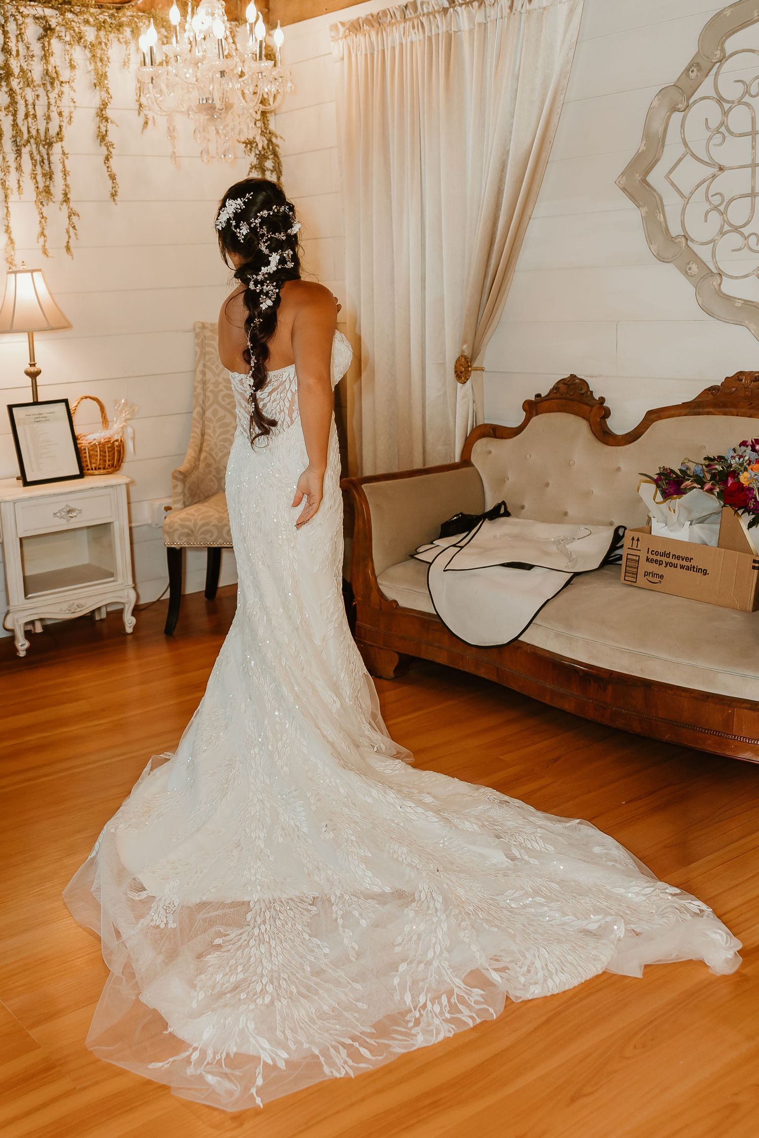 Bride in elegant lace mermaid wedding gown with long train, viewed from behind in a warmly decorated bridal suite.