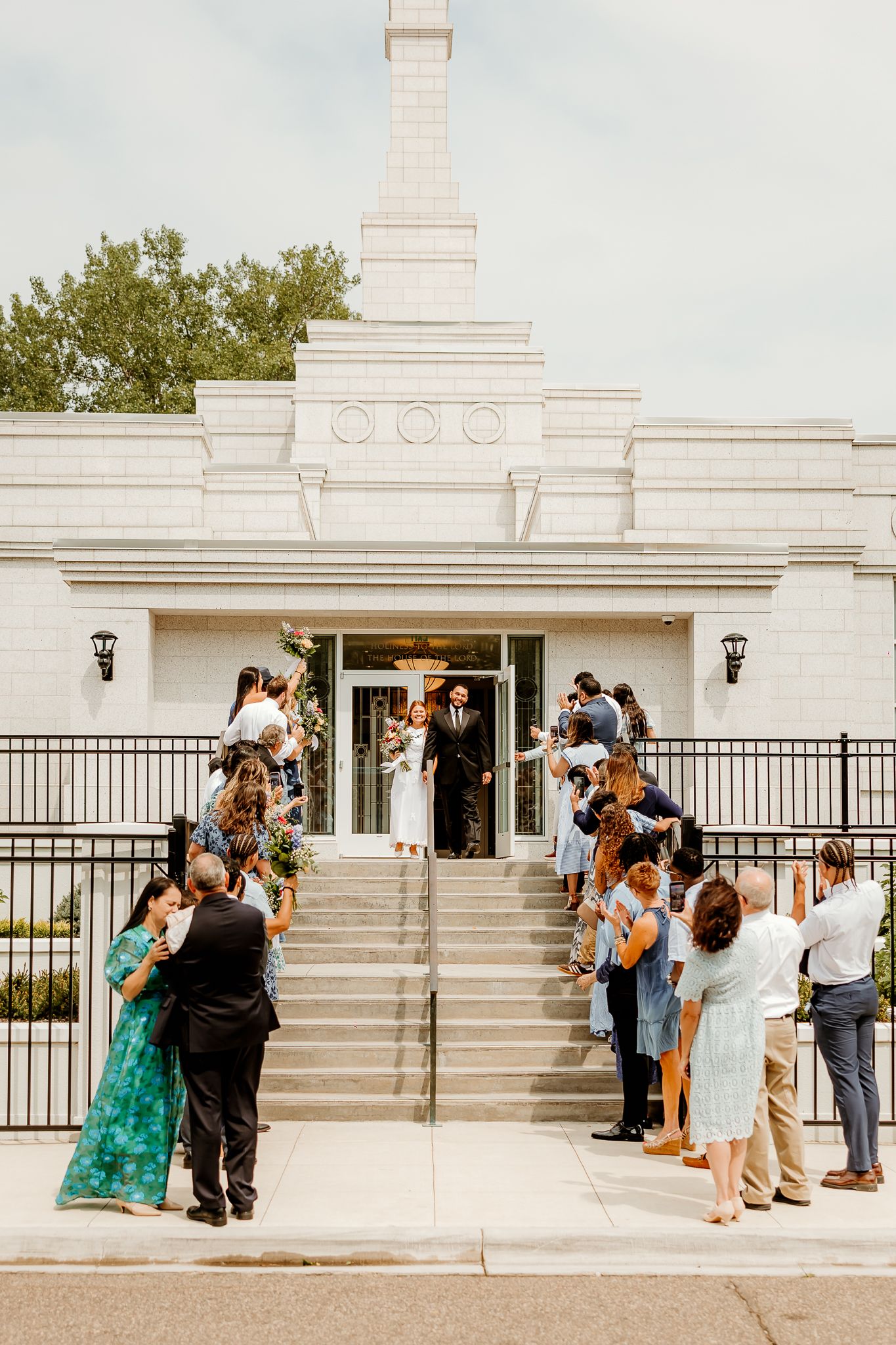 Wedding couple exits white LDS temple as guests celebrate on steps in sunny outdoor ceremony.