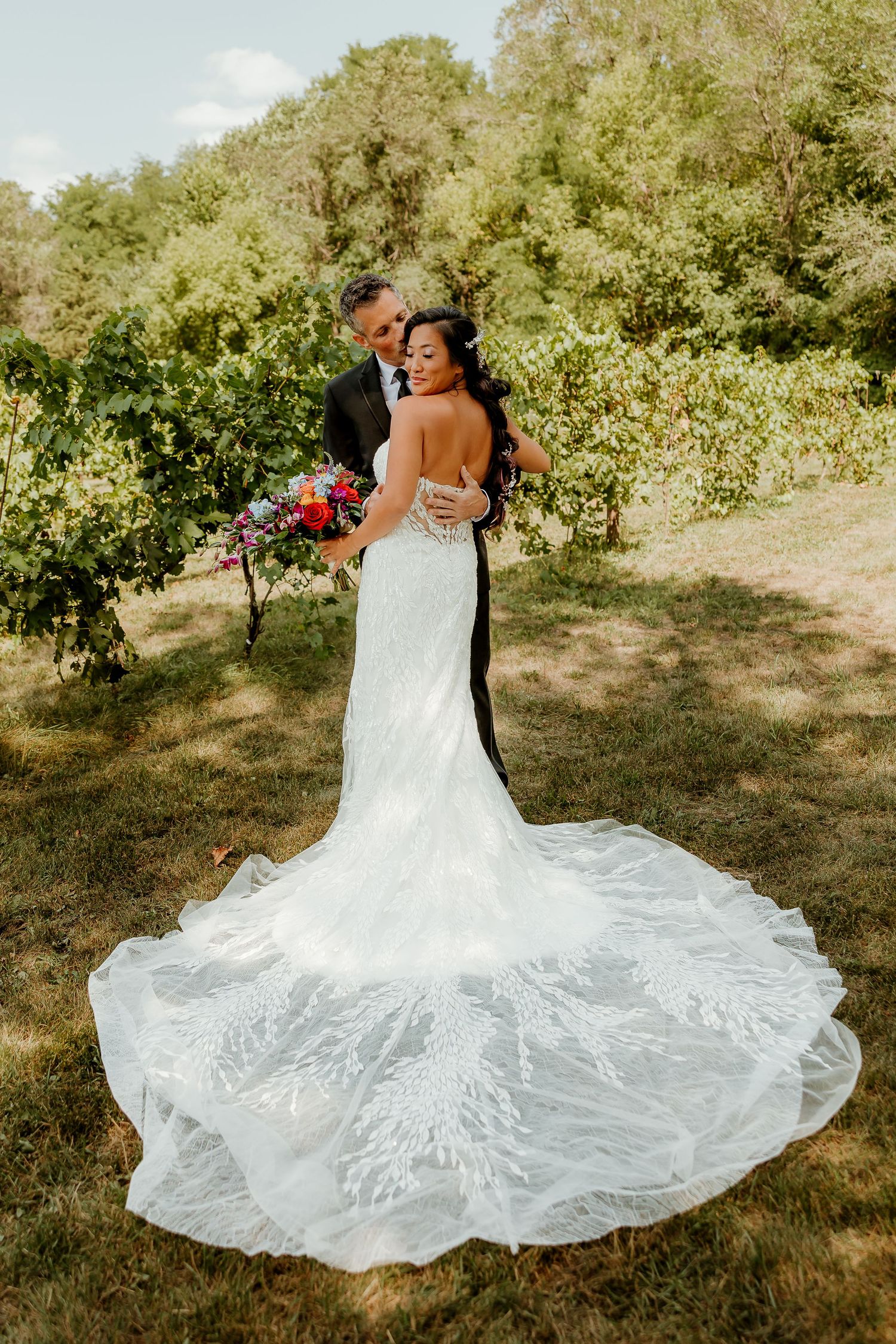 Bride in lace mermaid gown with train embraced by groom in tuxedo, posing in lush green vineyard setting.