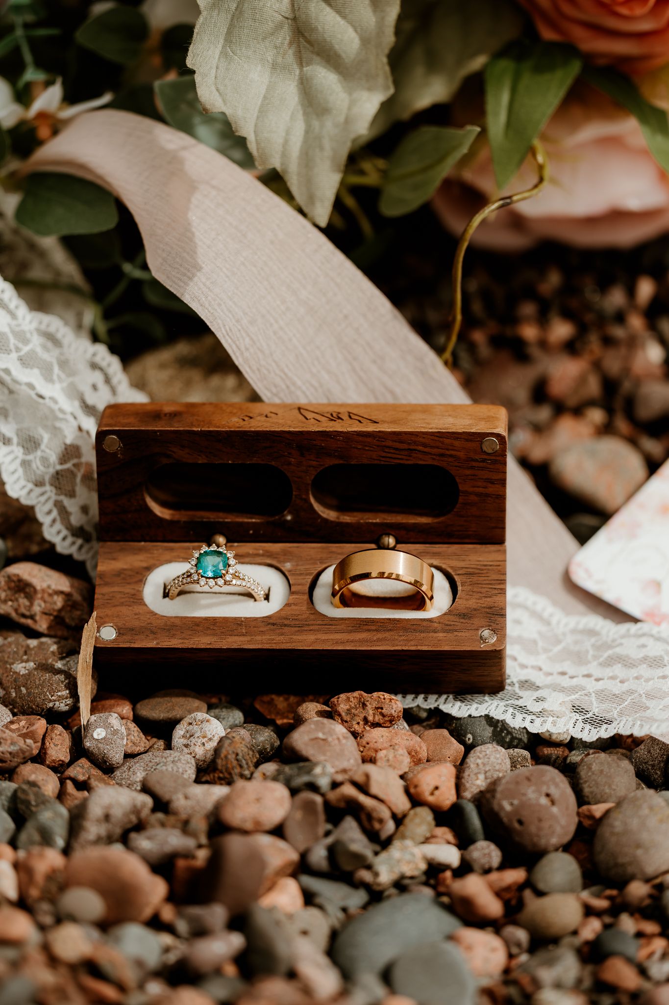 Two gold wedding rings, one with emerald stone, displayed in a wooden ring box on pebbles with floral backdrop.