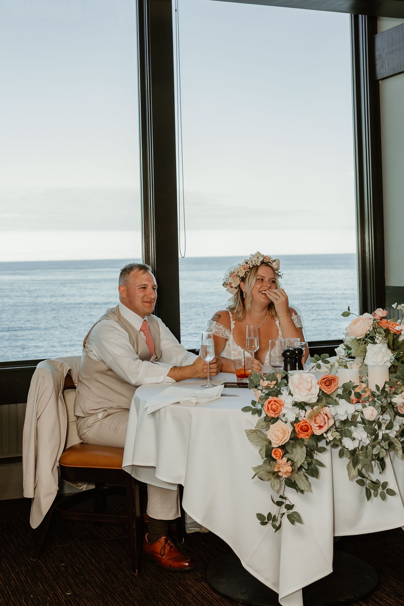 Bride and groom seated at floral-decorated sweetheart table with ocean view backdrop at coastal wedding reception.