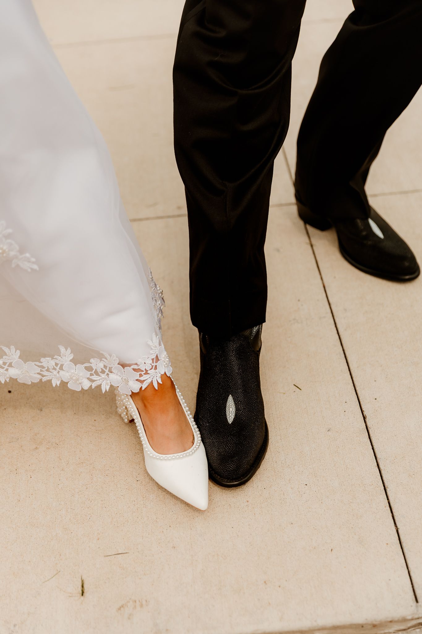 Close-up of bride's white pointed-toe heels beside groom's black cowboy boots on stone pavement at wedding.