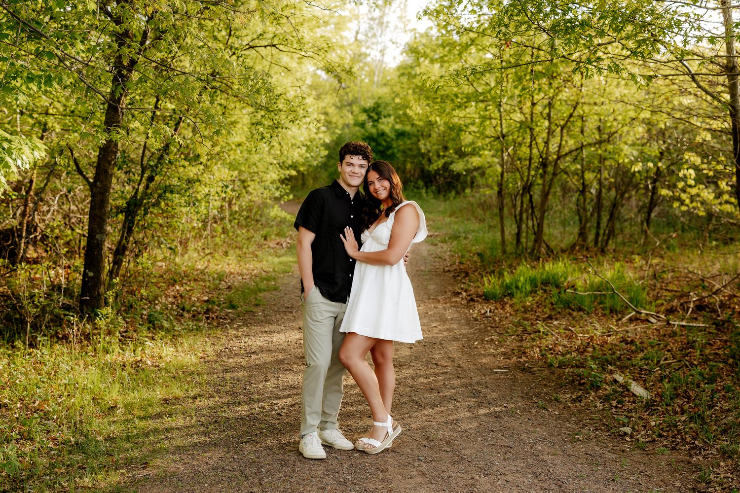 A couple stands together on a sunlit forest path, the woman in a white dress leaning on the man.