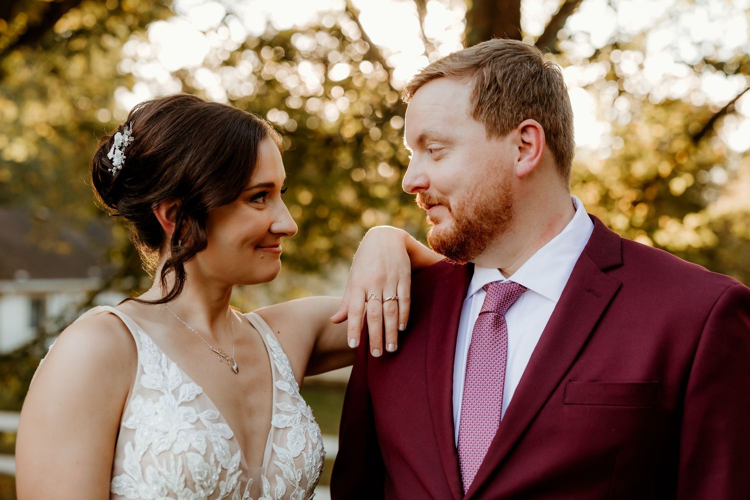 A smiling bride in a lace gown with a hair accessory rests her hand on the shoulder of a groom in a burgundy suit and pink tie outdoors.