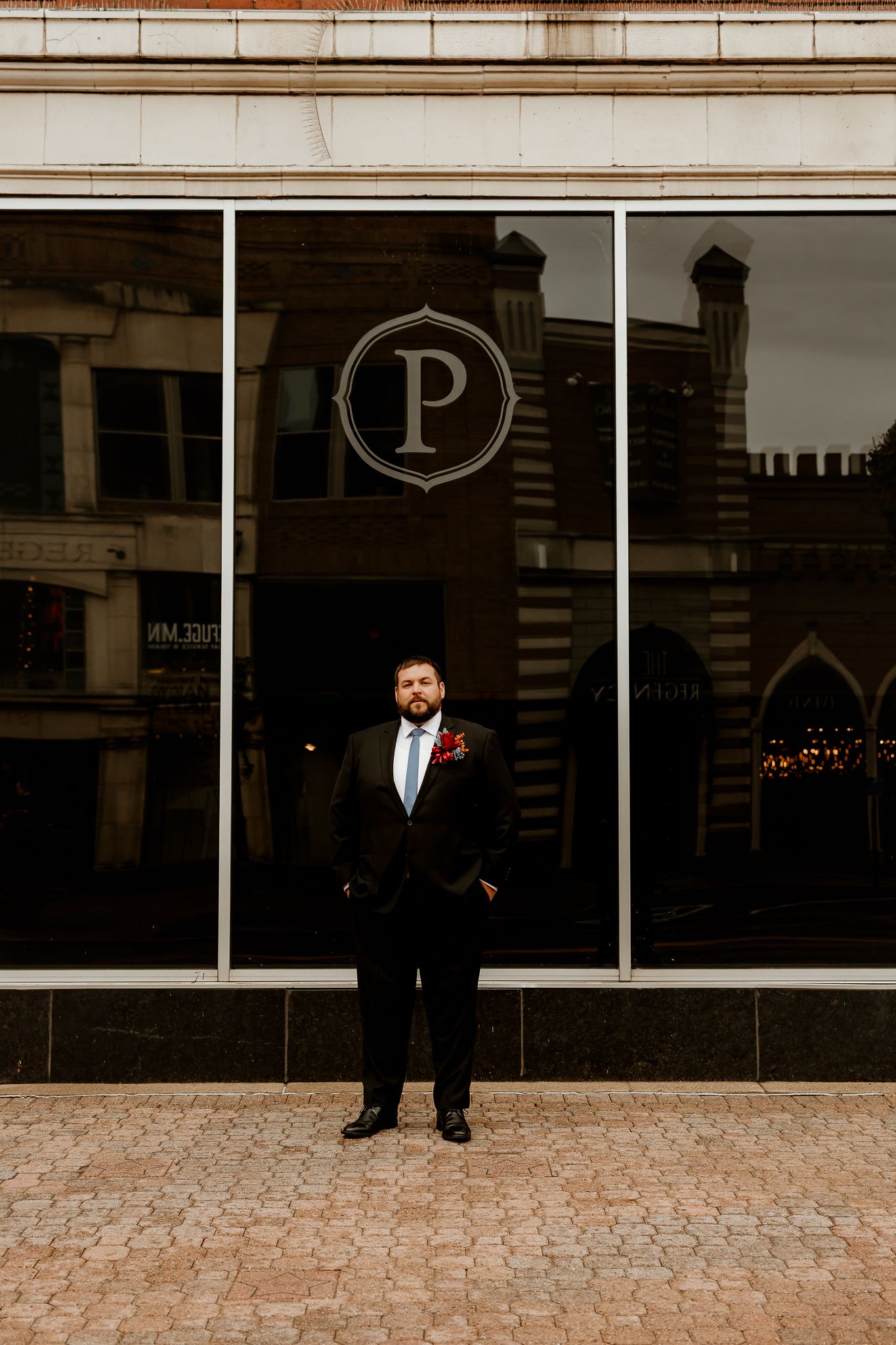 Groom in a dark suit, light blue tie and red boutonniere stands on a brick sidewalk before a glass storefront with a large P logo.