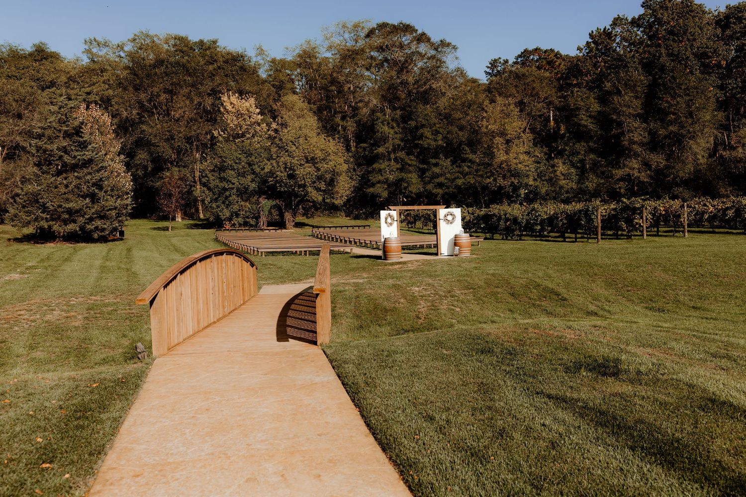 Curved wooden footbridge over a grassy lawn leading to a small white ceremony arch, with a vineyard and tall green trees in the background.