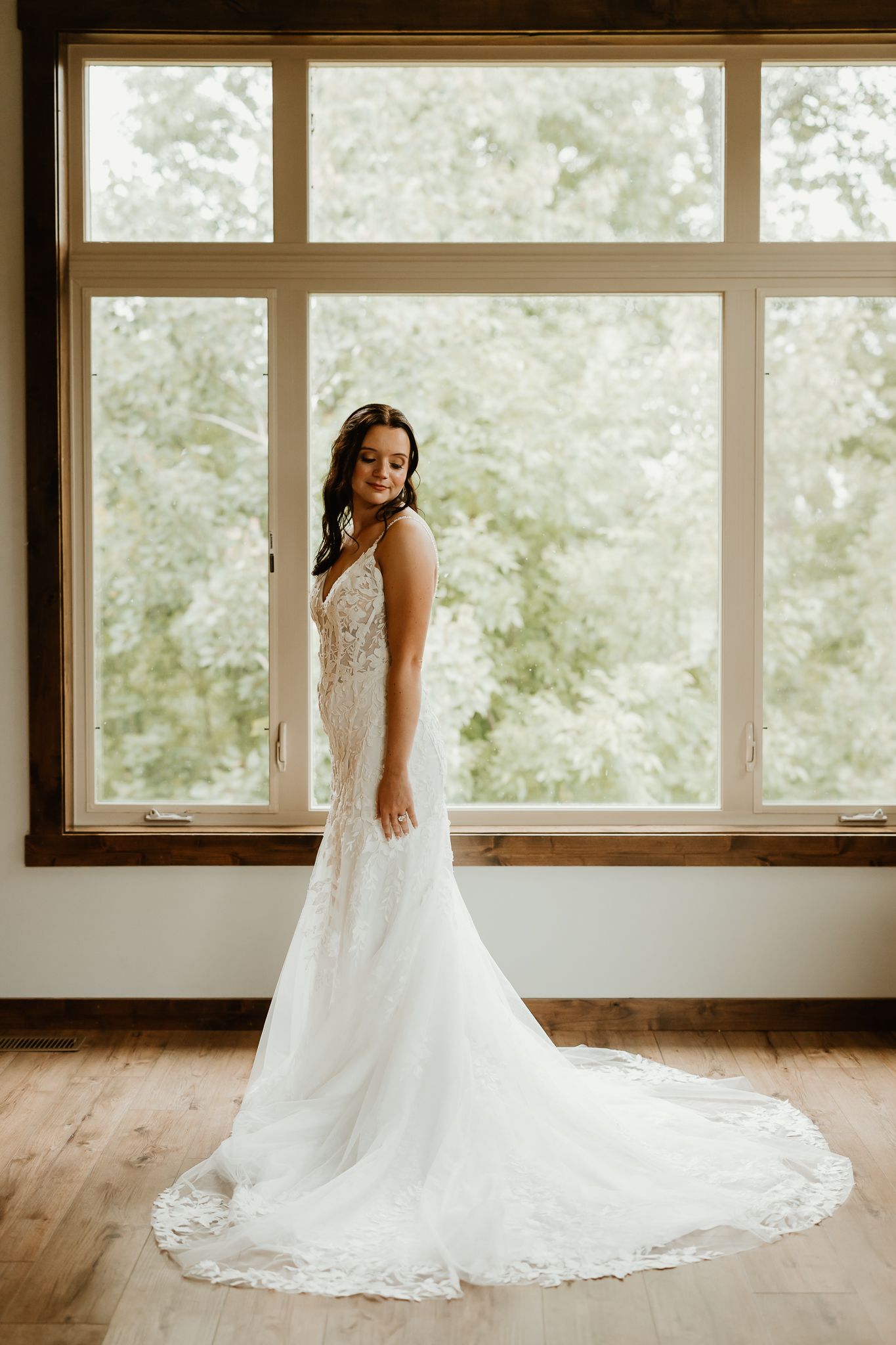 Bride in an elegant white lace wedding gown with a long train stands beside a large window overlooking green trees.