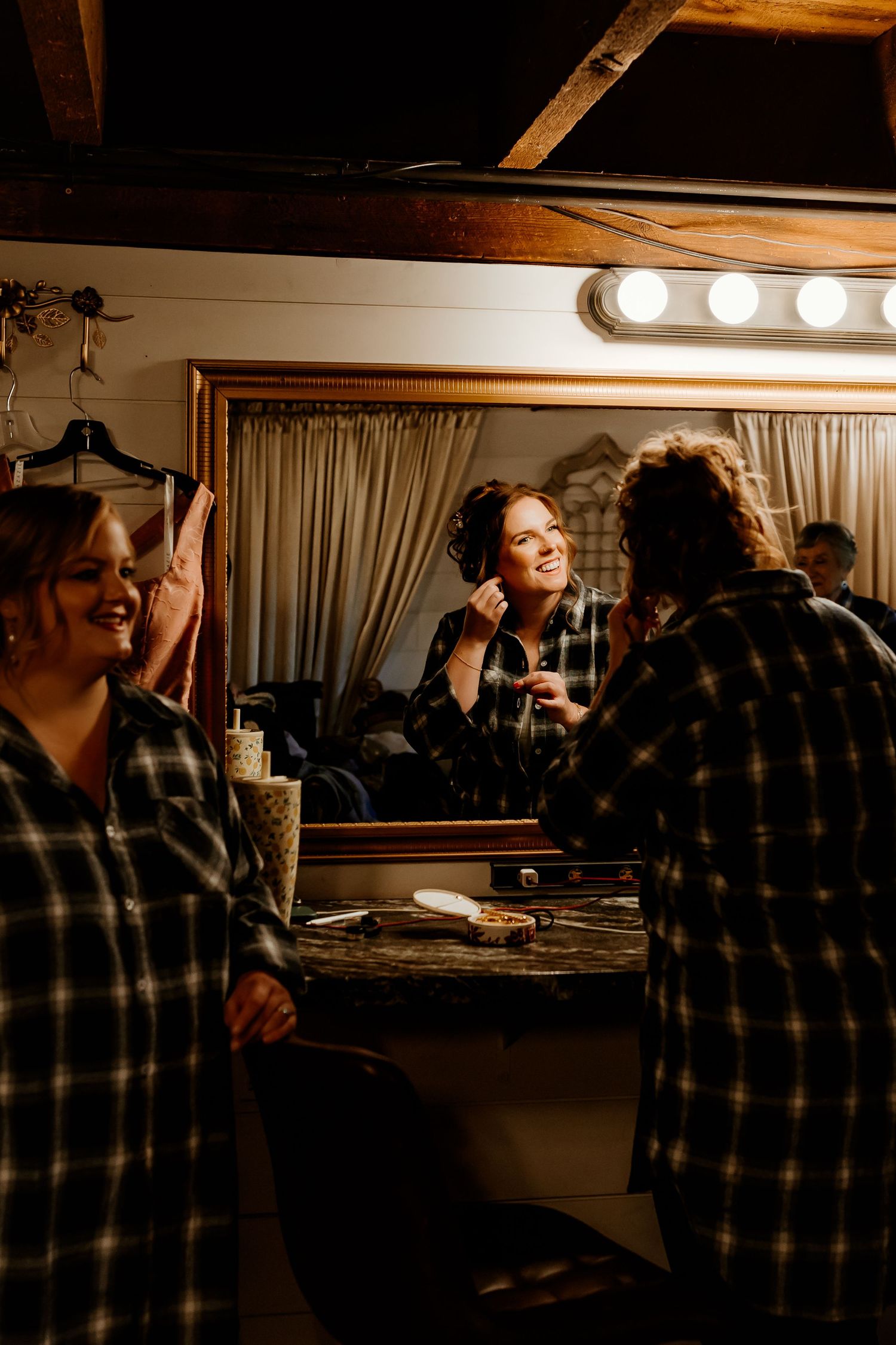 Women in matching plaid flannel shirts getting ready in front of a vanity mirror with bright bulbs, one smiling while applying makeup.