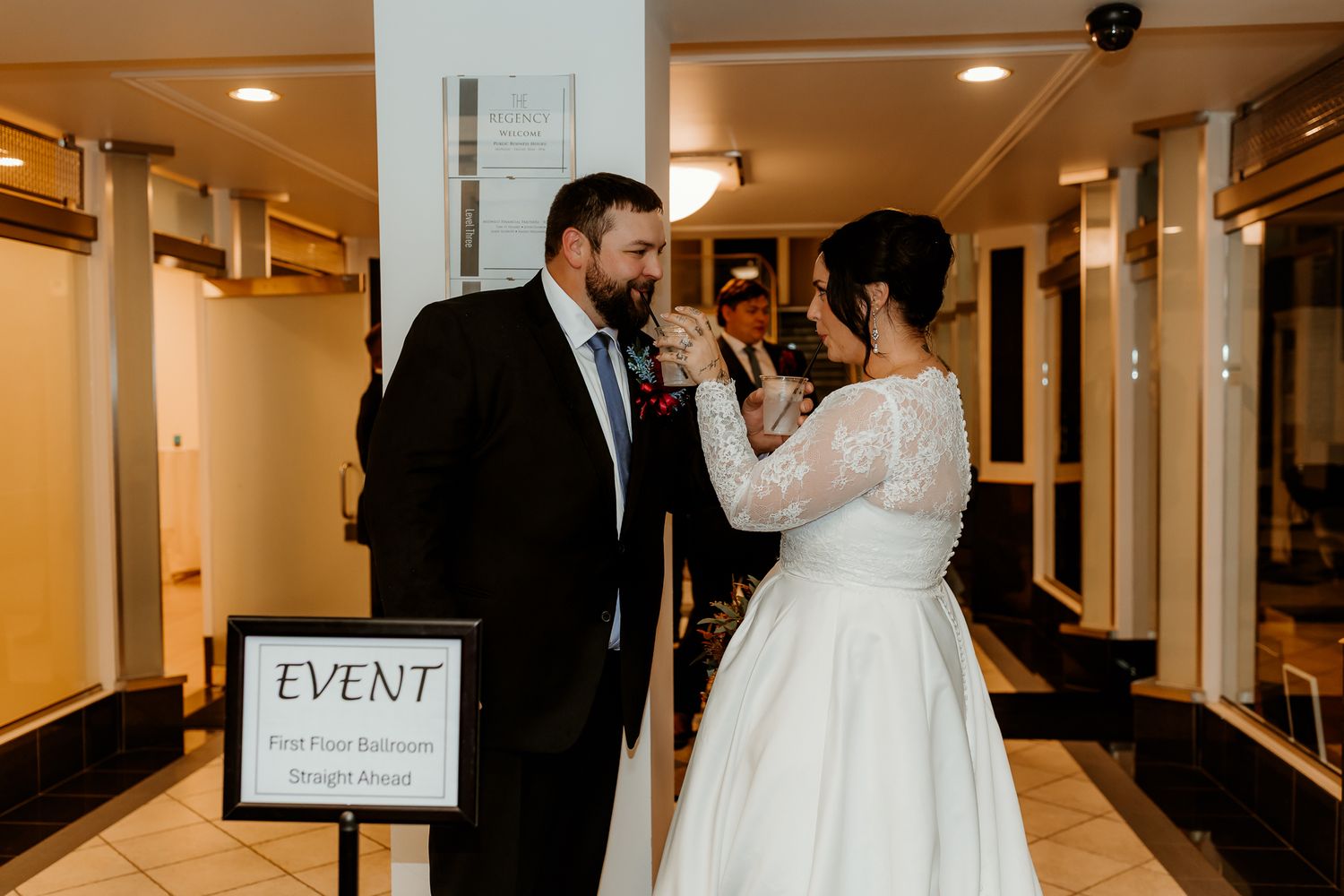 Bride in a white lace gown and groom in a dark suit share a toast in a hotel hallway beside an event sign.