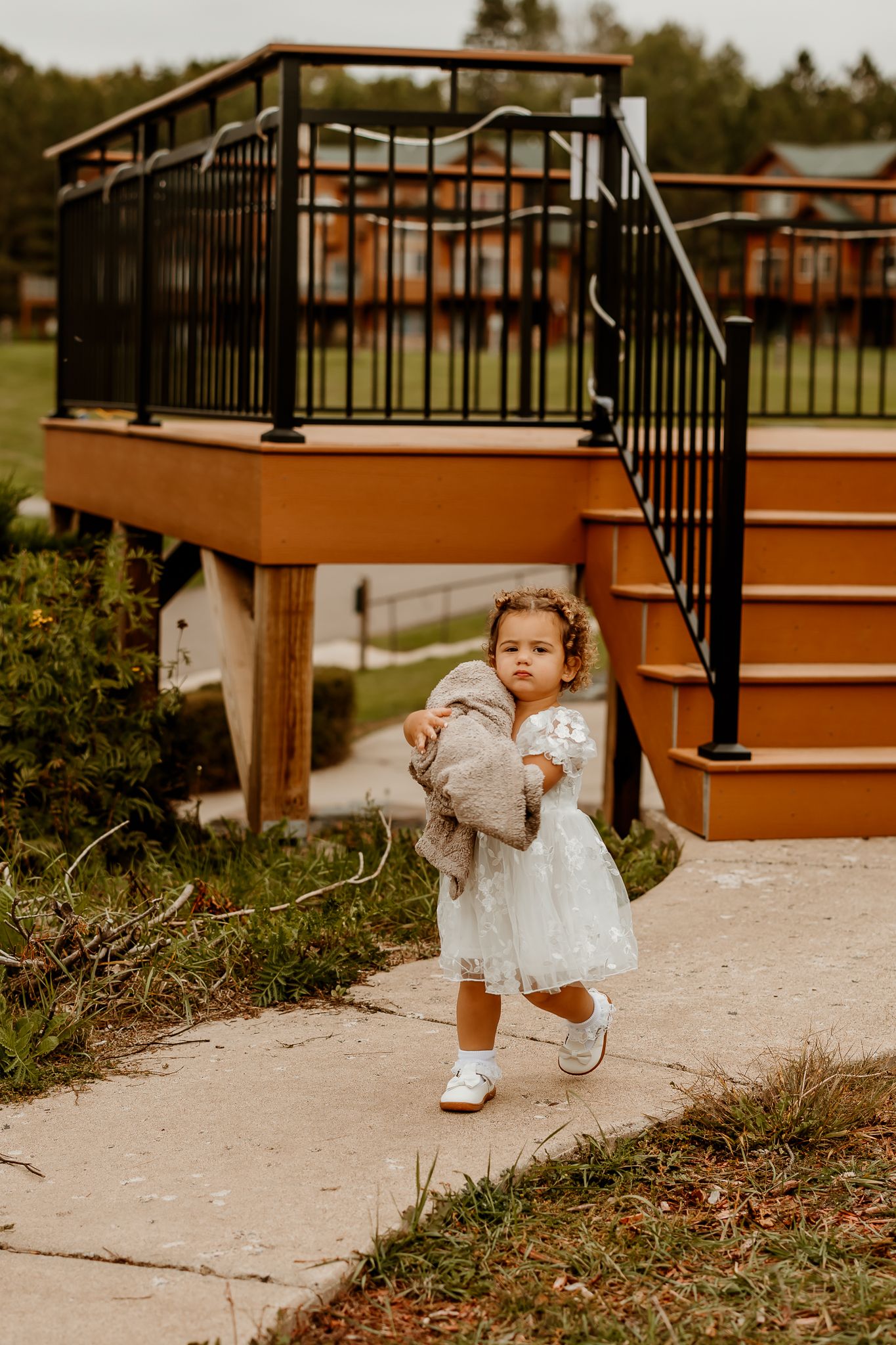 Toddler in a white lace dress and white shoes carries a gray coat along a concrete path beside a wooden deck with black railings.