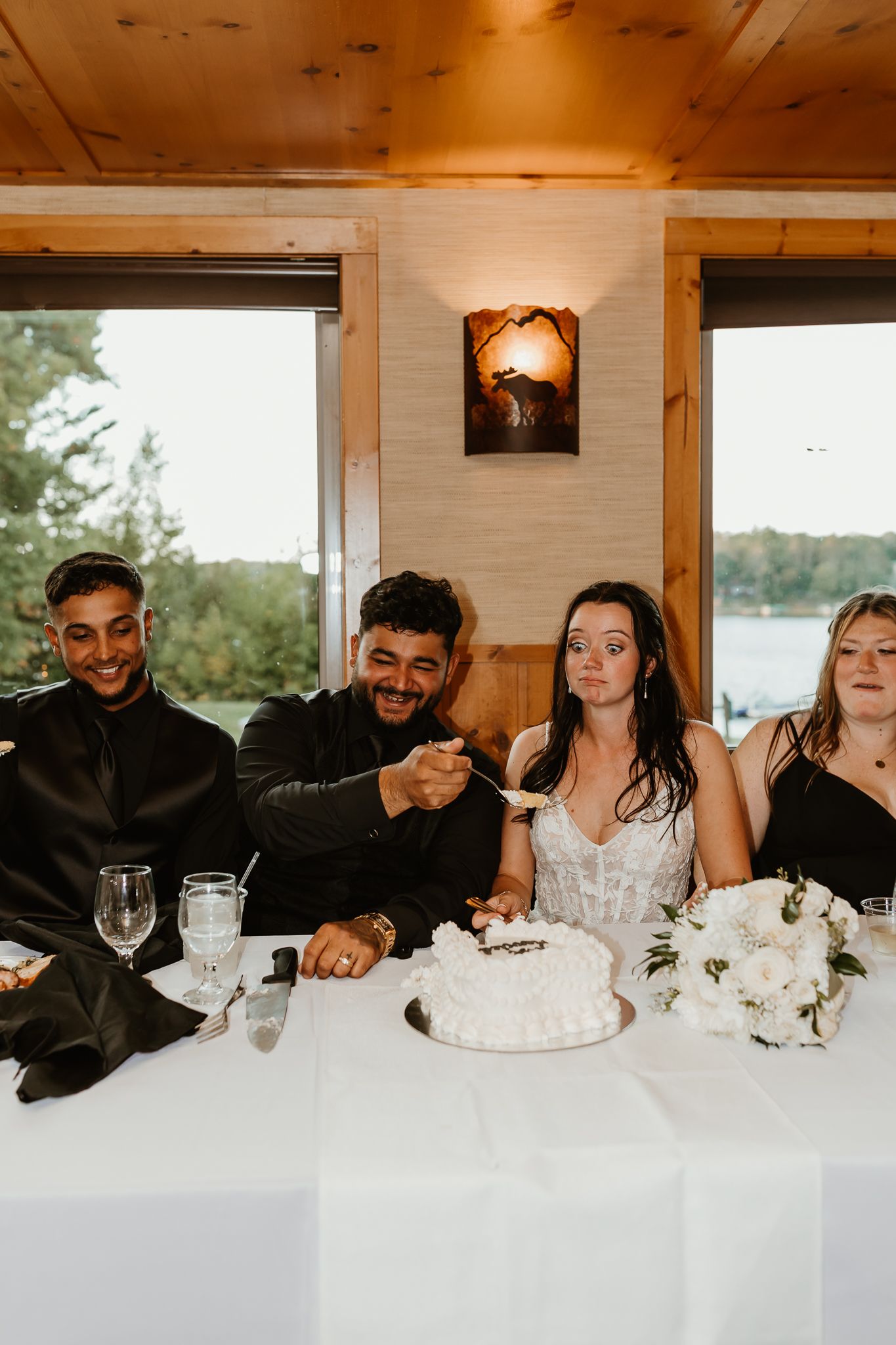 Wedding party seated at a white-draped table with a small white cake and bouquet, as a groom in black laughs while feeding a bite of cake.