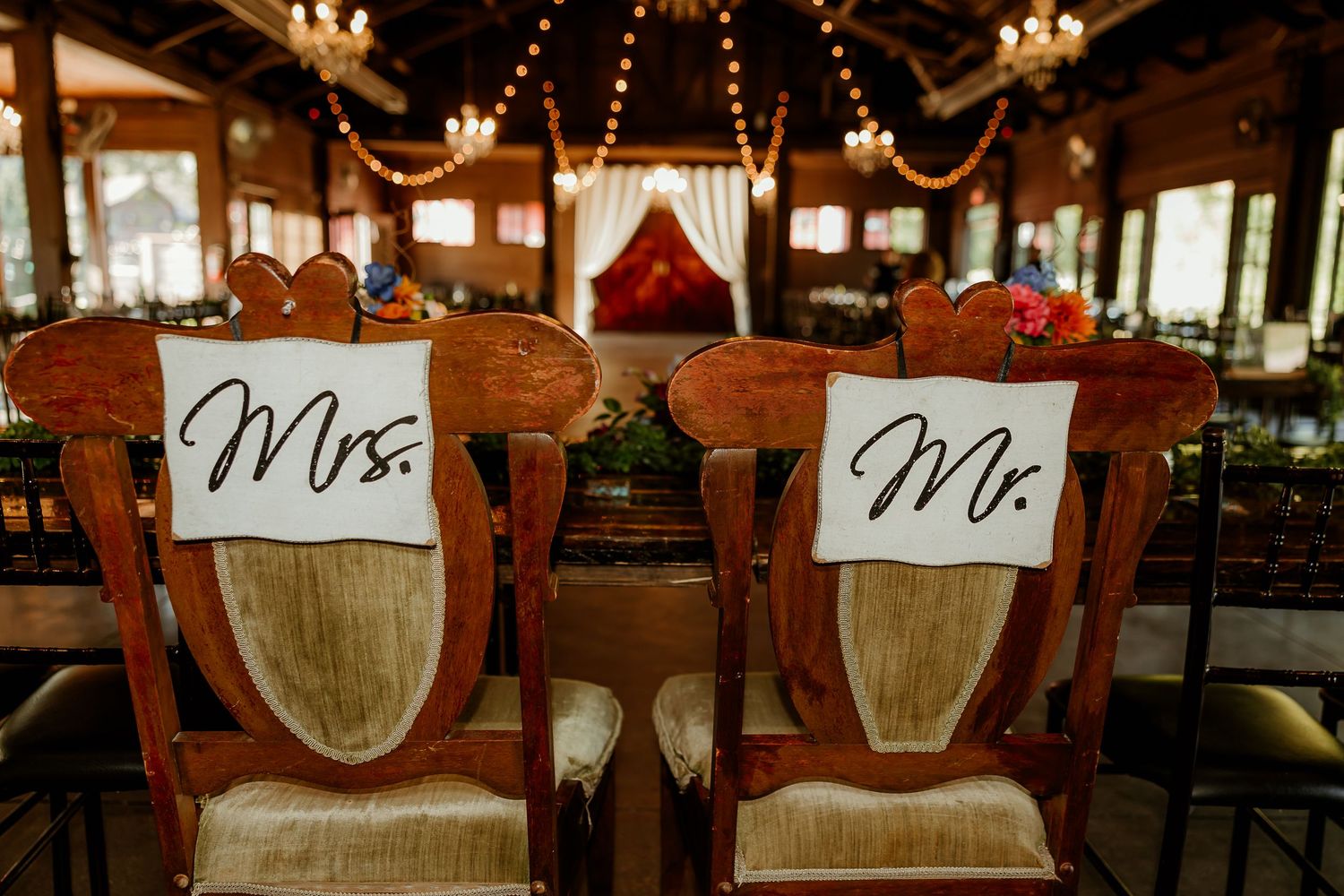 Mr. and Mrs. signs hang on ornate wooden wedding chairs in a rustic reception venue with warm string lights.
