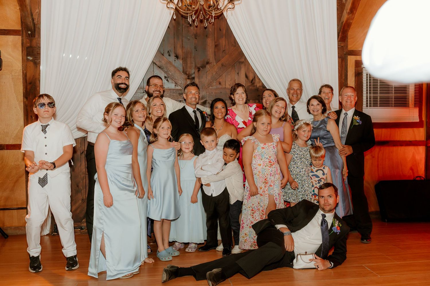 Large wedding family group photo with adults and children posing together in elegant venue with chandelier.