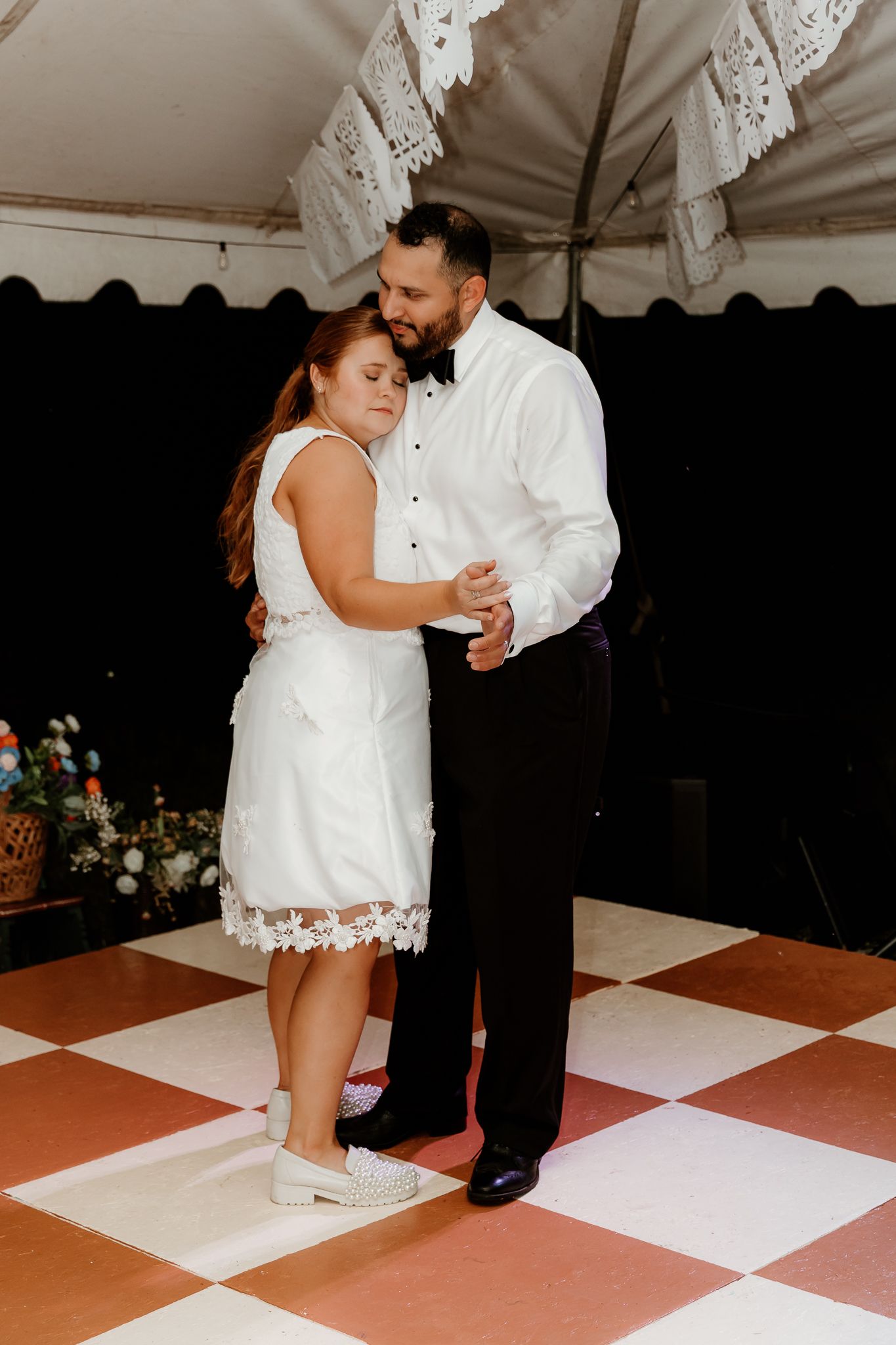 Bride and groom share their first dance on a checkered floor under a white tent at their wedding reception.
