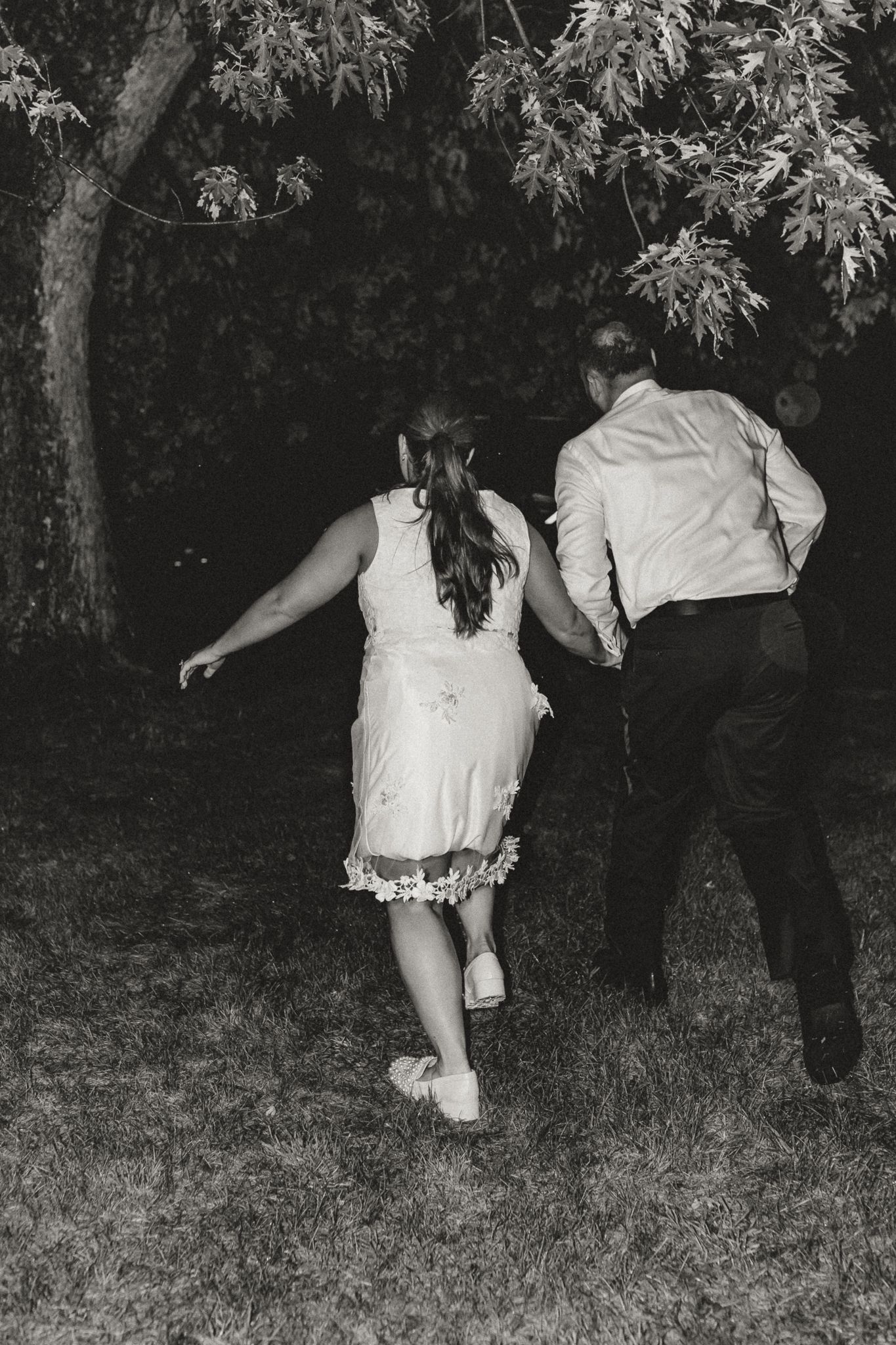 Black and white photo of a couple holding hands running across a grassy lawn at night near trees.