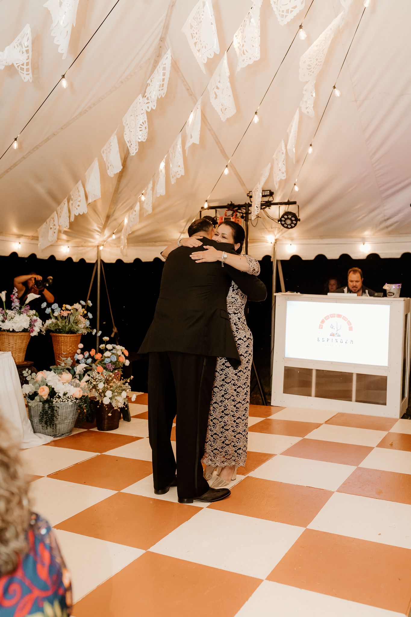 Couple sharing a tender dance embrace on checkered dance floor inside decorated wedding tent with string lights.