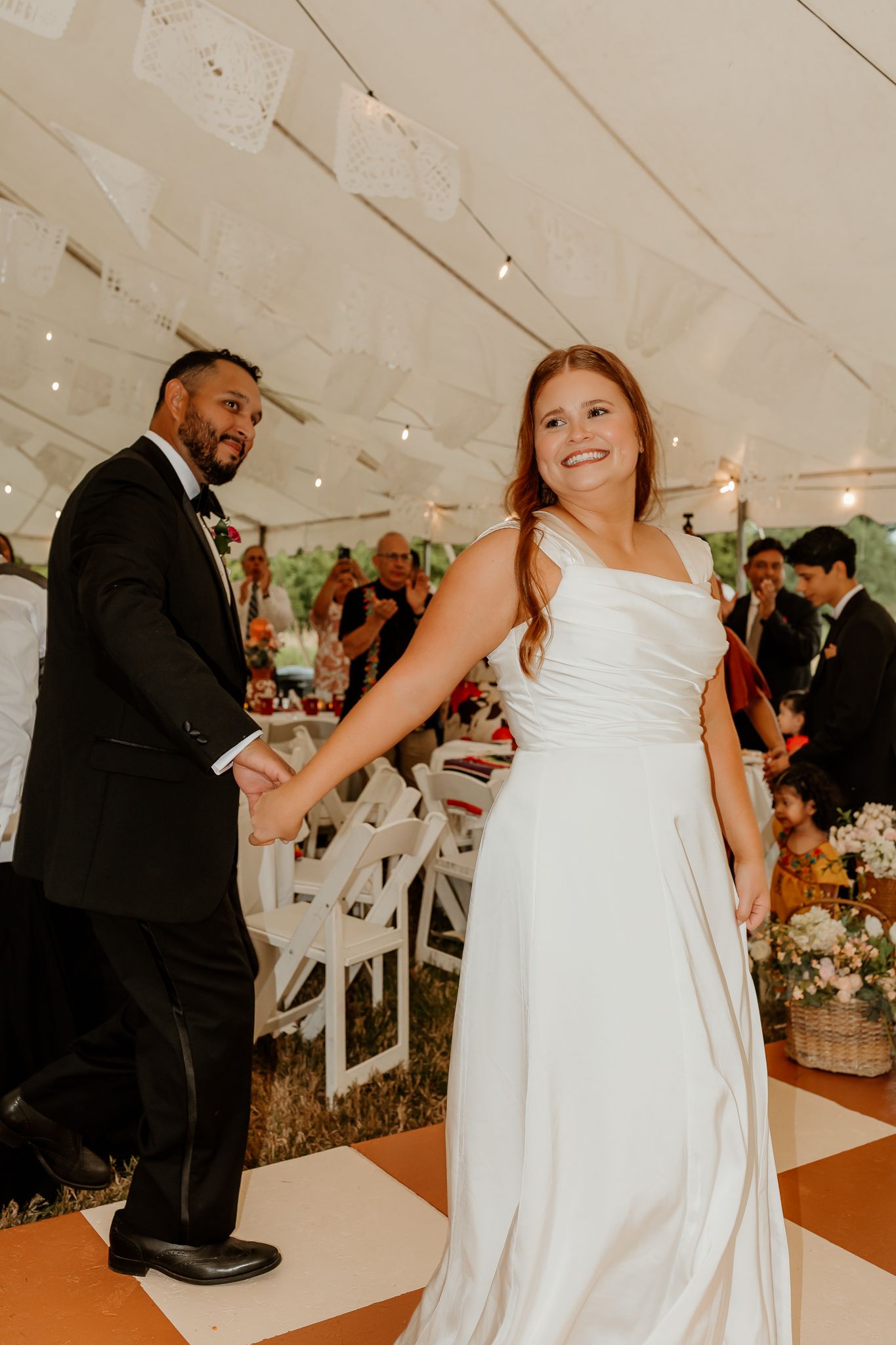 Bride in white gown and groom in black tuxedo hold hands while entering their wedding reception under a tent.