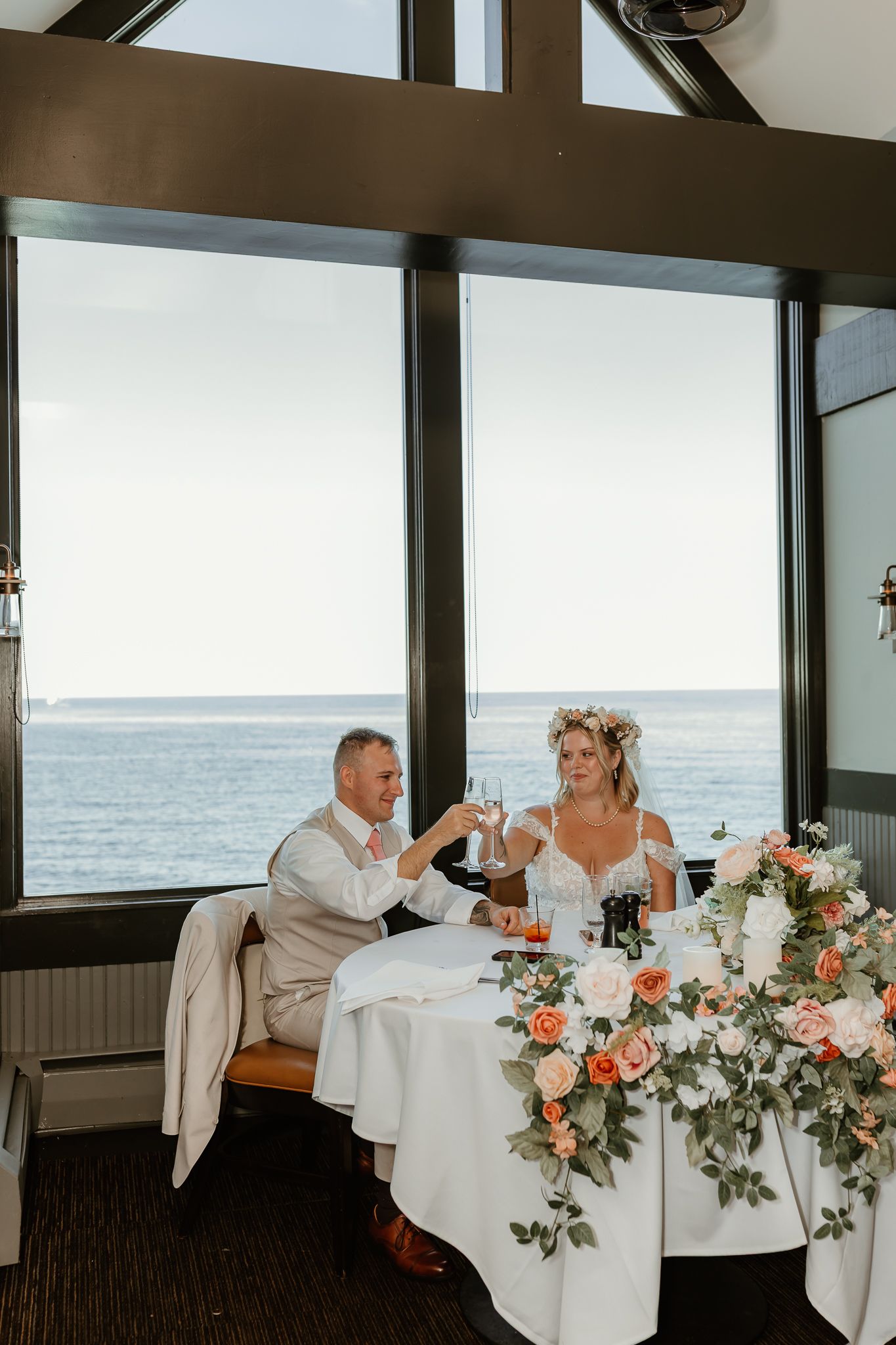 Bride and groom toasting champagne at sweetheart table with ocean view and floral arrangements at seaside wedding reception.