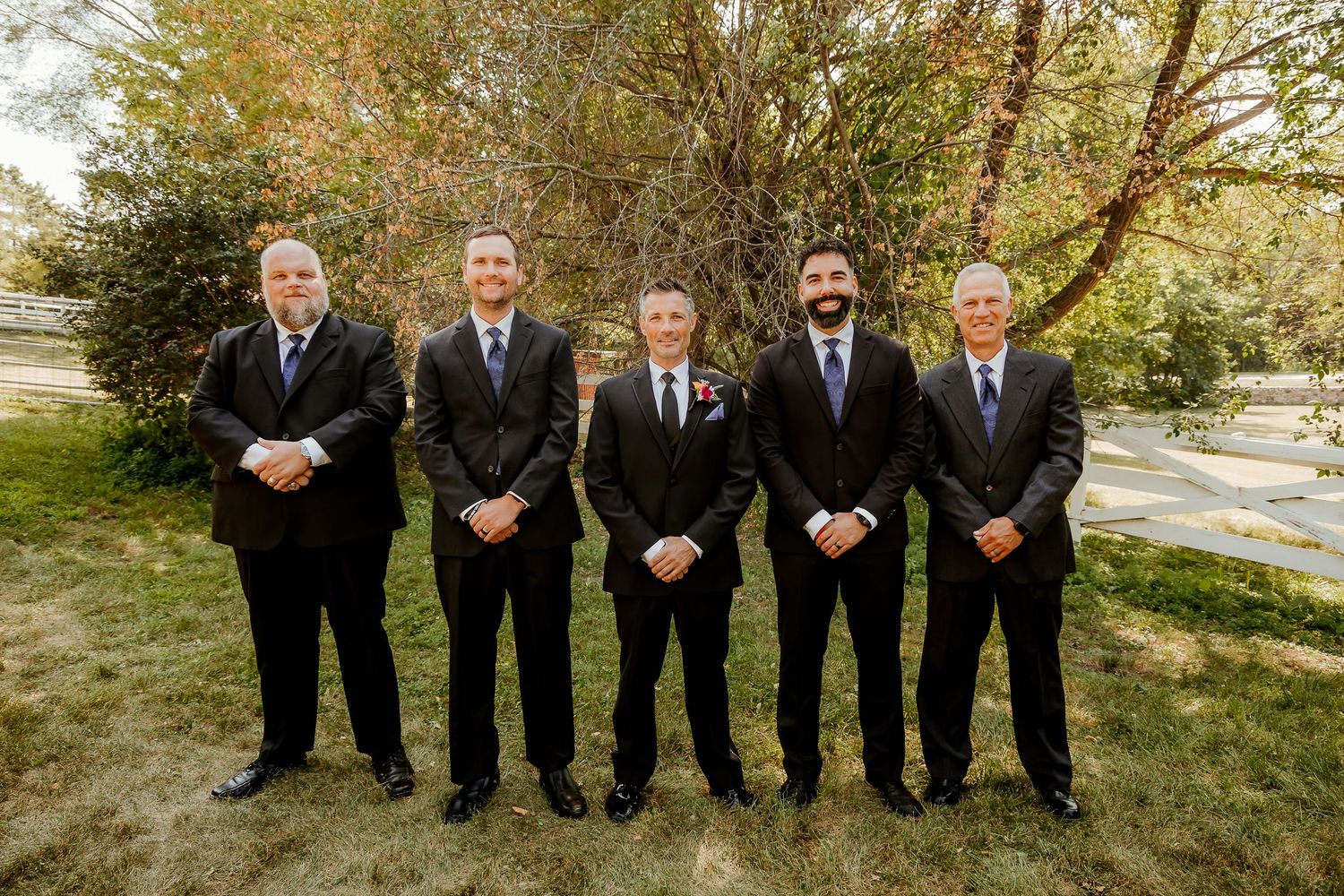 Five groomsmen in black suits and ties posing outdoors on green grass with trees in the background at a wedding.