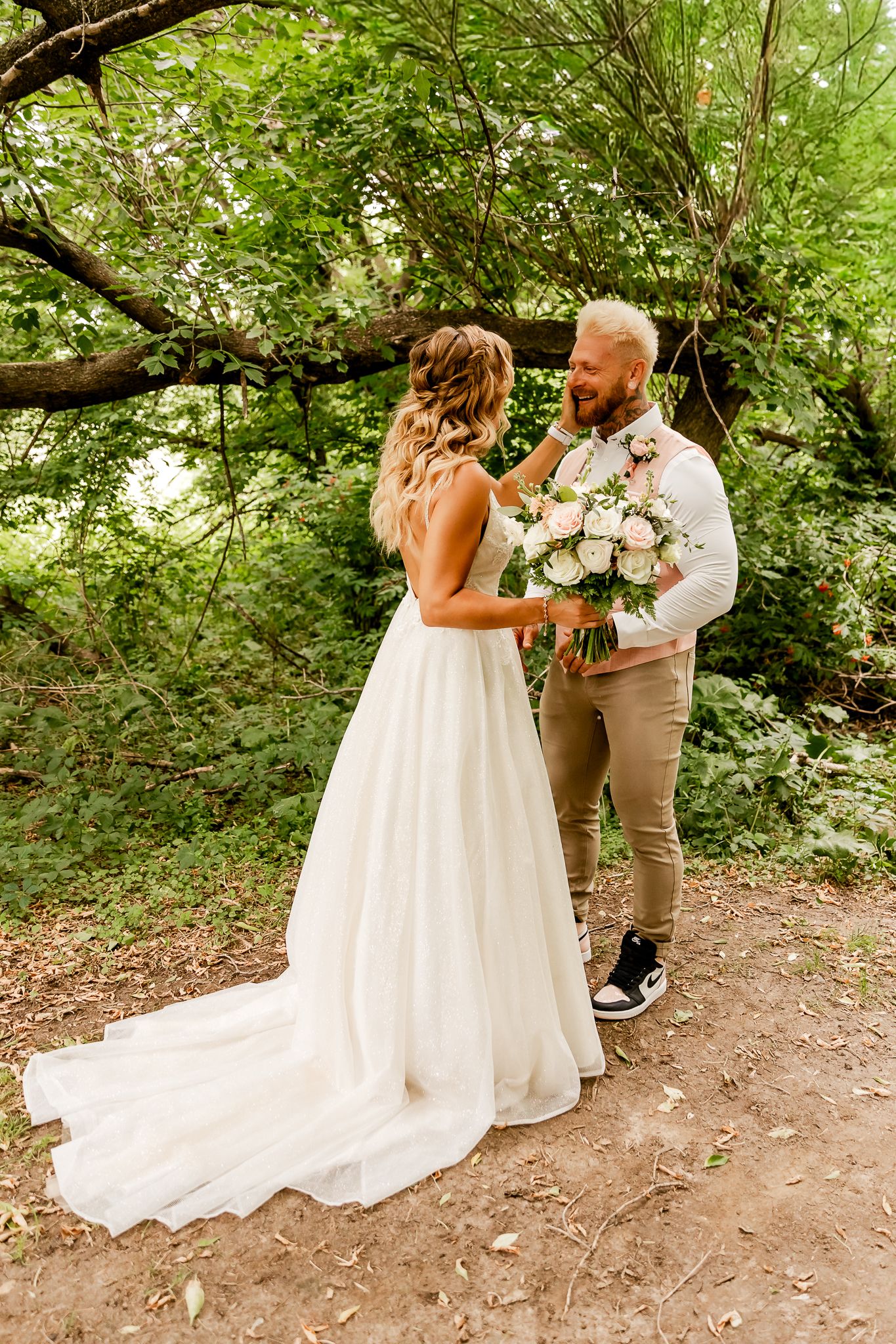 A bride in a white gown and groom in khakis share a joyful moment with a pink bouquet in a lush green forest.