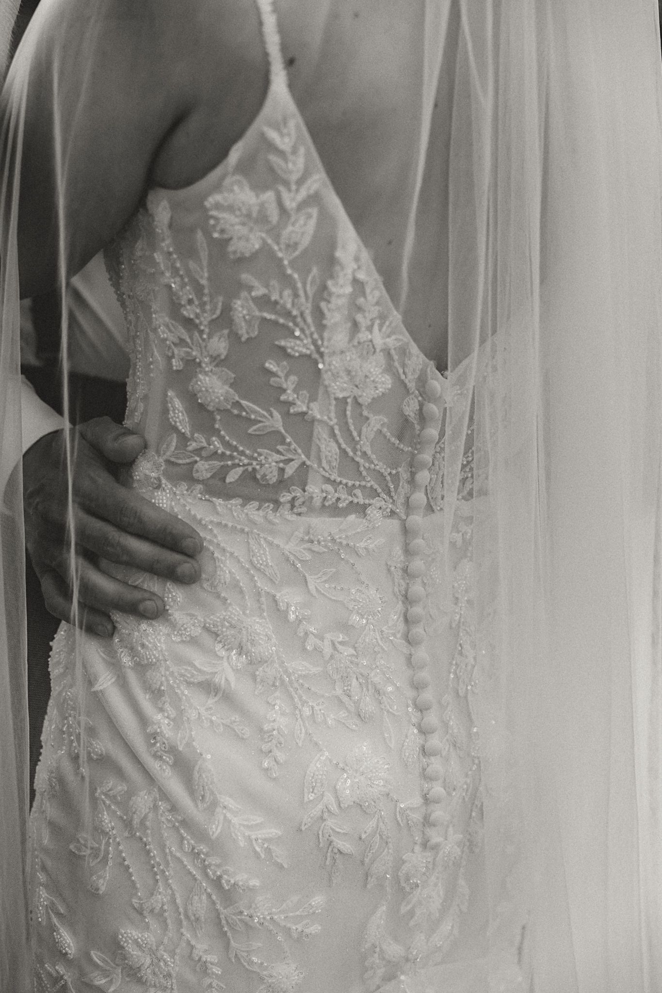 Black and white photo of a bride in an intricate lace wedding gown with floral embroidery and a flowing veil.