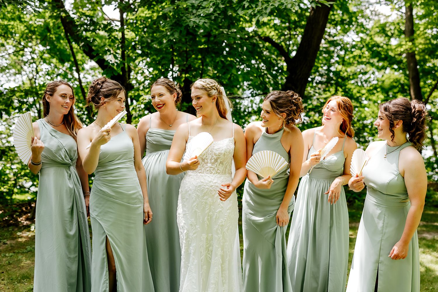 Bride in white lace gown stands with six bridesmaids in sage green dresses, laughing outdoors among lush green trees.
