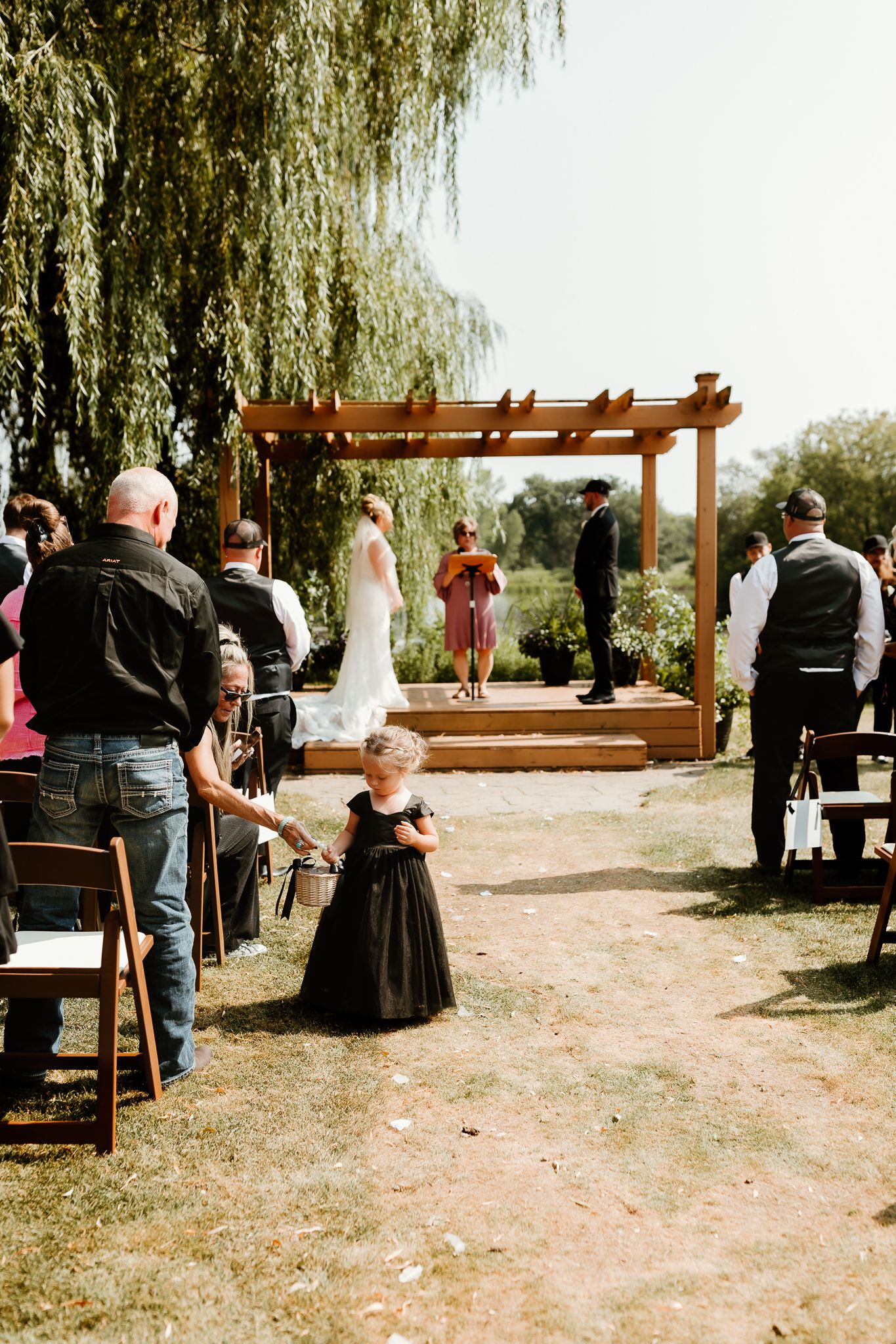 Outdoor wedding ceremony with flower girl in black dress walking down aisle toward pergola altar under willow tree.