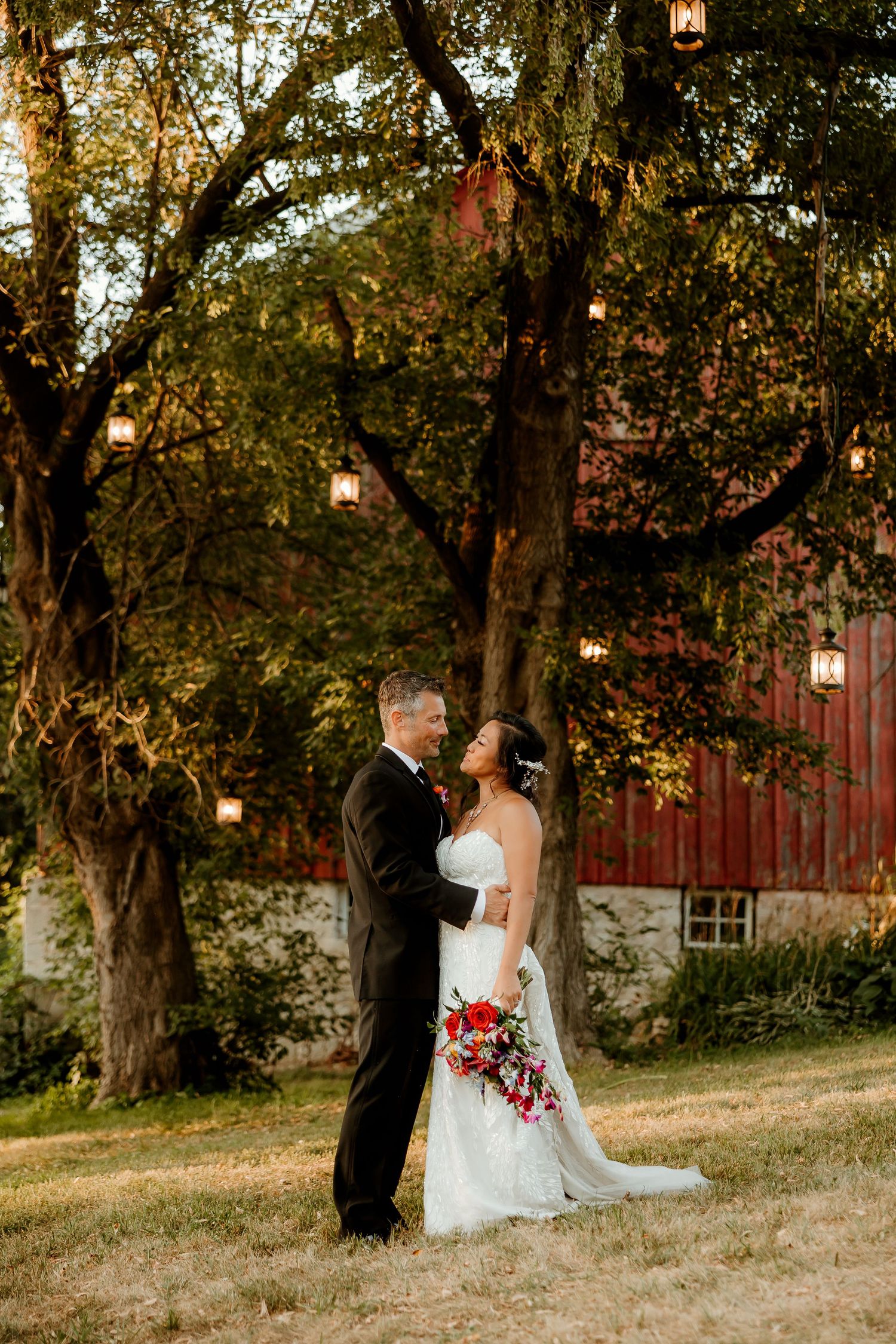 Bride and groom embrace under lantern-lit trees beside a red barn at golden hour wedding.