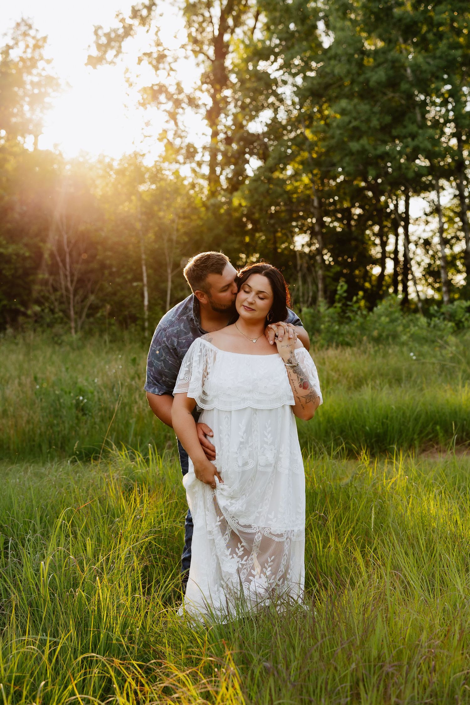 Couple embracing at golden hour in green field, man kissing woman in white lace boho dress.