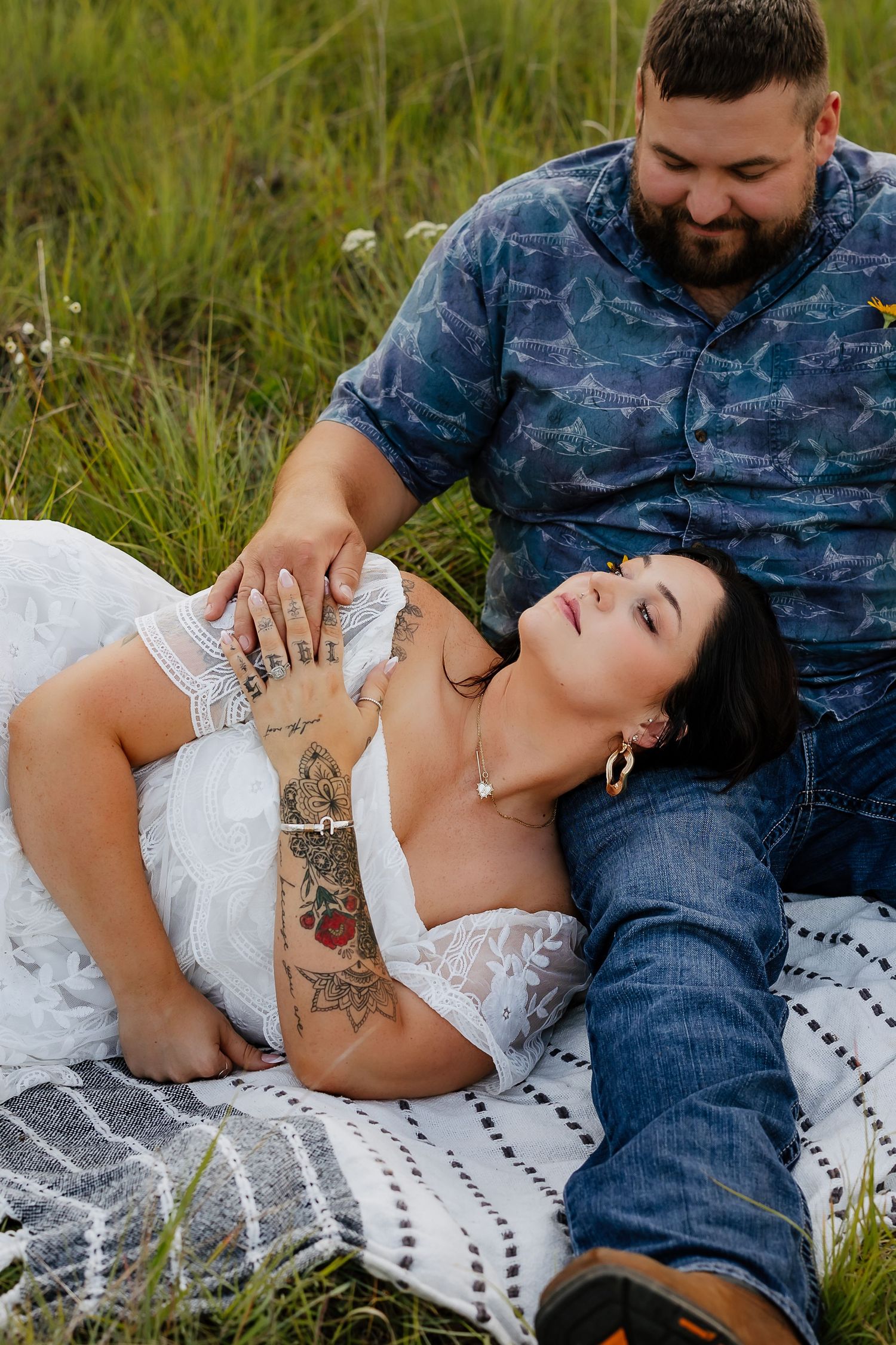 Couple relaxing on blanket in tall grass, tattooed woman in white dress lying across mans lap smiling.