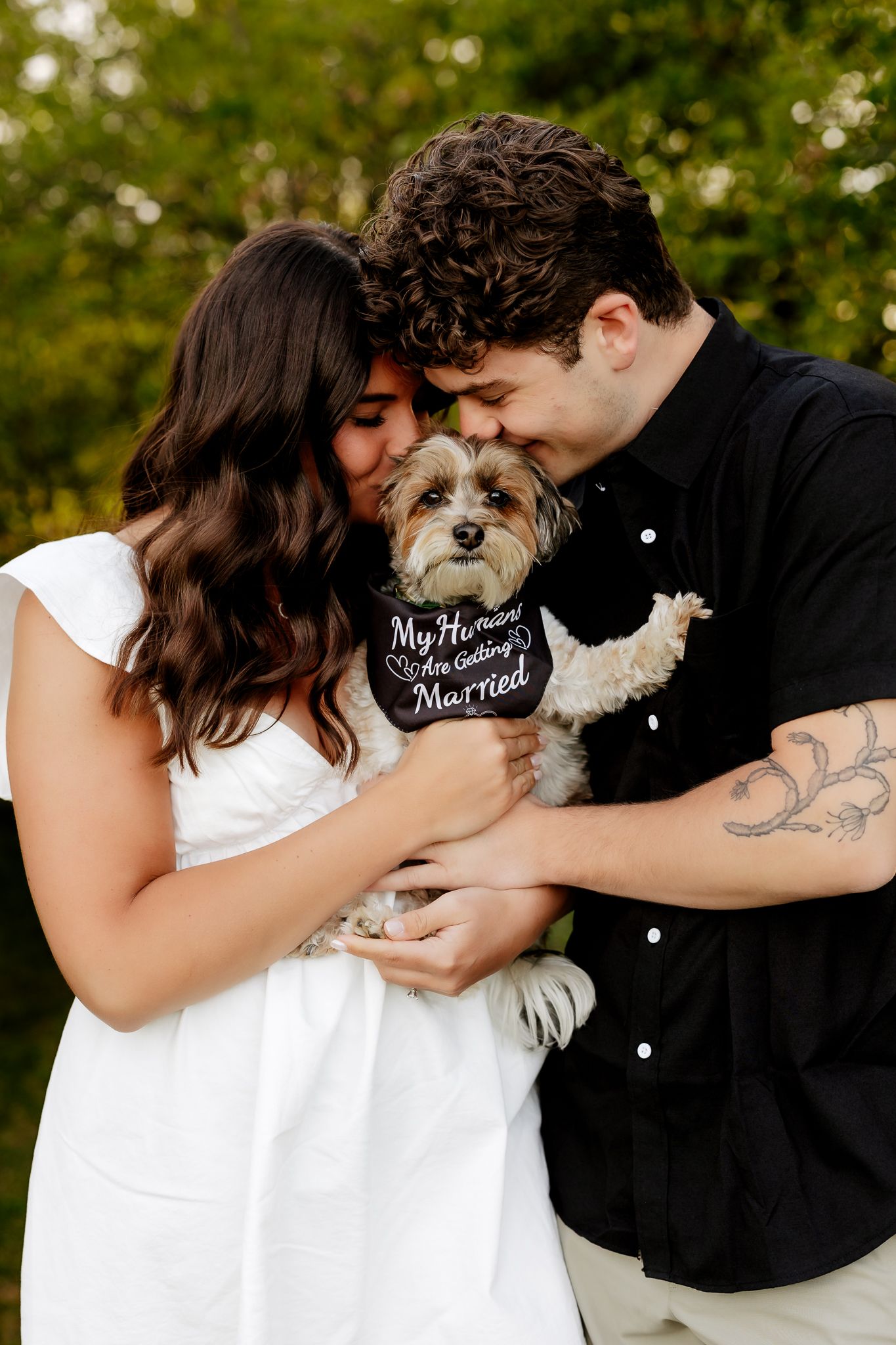 An engaged couple leans in to kiss their small dog wearing a My Humans Are Getting Married bandana outdoors.