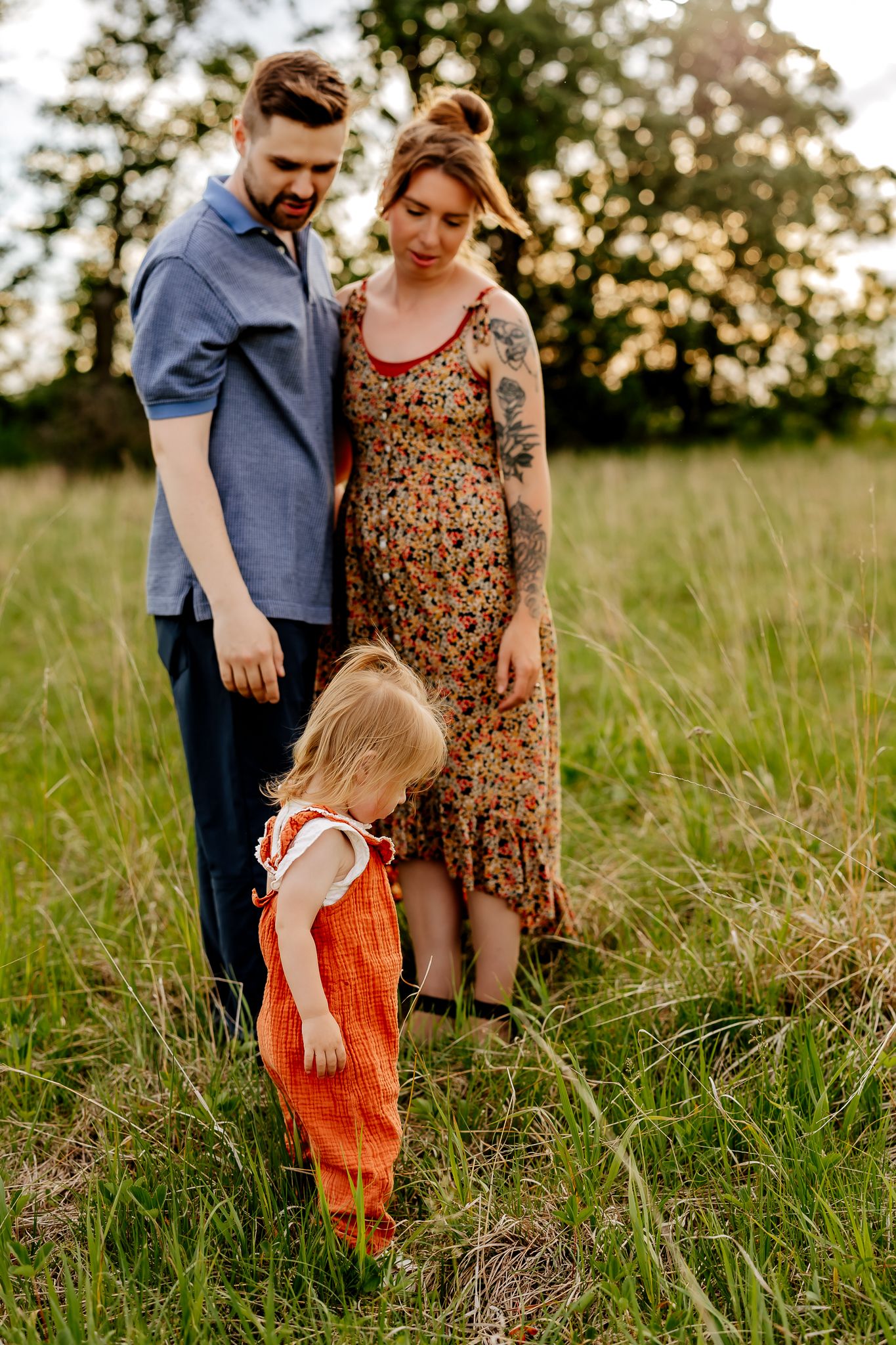 A couple watches their young toddler in an orange outfit explore a lush green meadow at sunset.