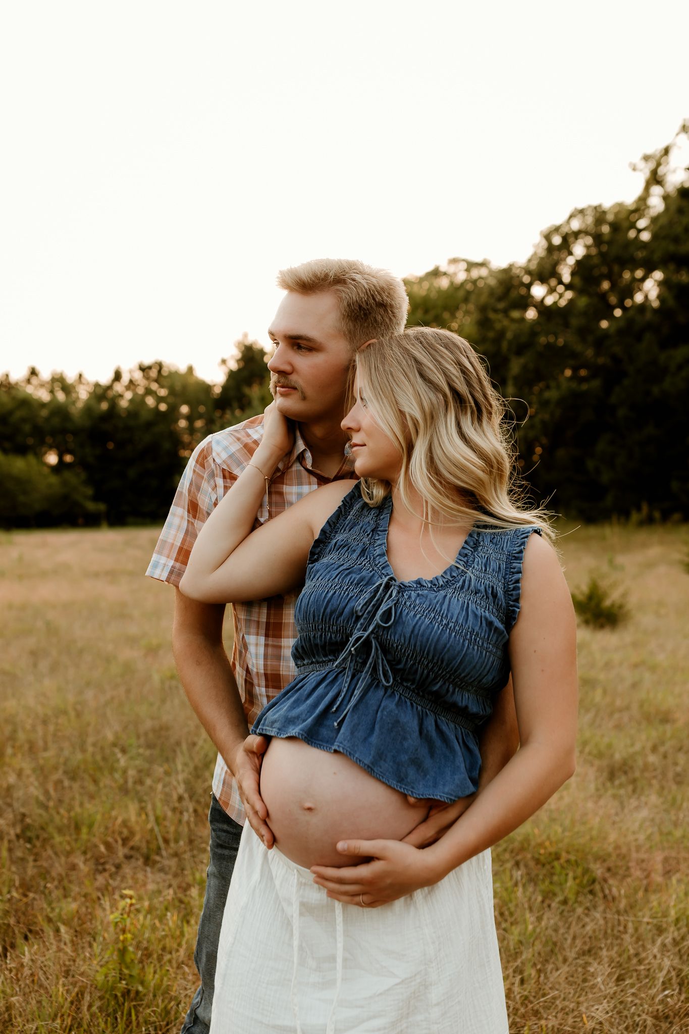Couple in a golden field, pregnant woman in denim top and white skirt, partner embracing her from behind at sunset.