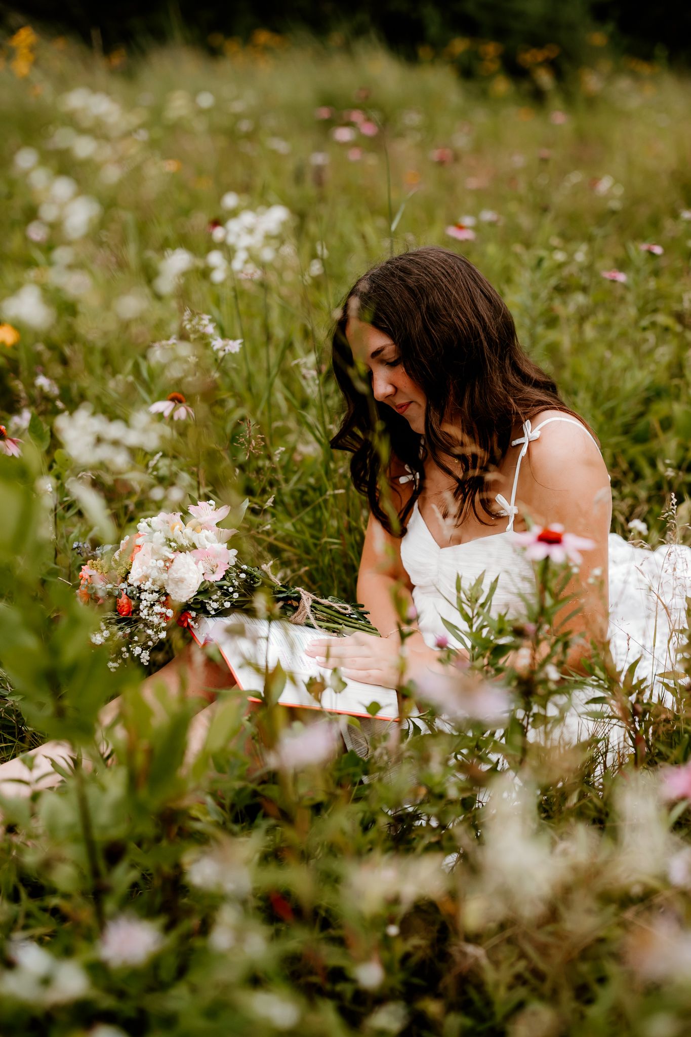 A brunette bride in a white dress sits in a lush wildflower meadow holding a bouquet of pink and white flowers.