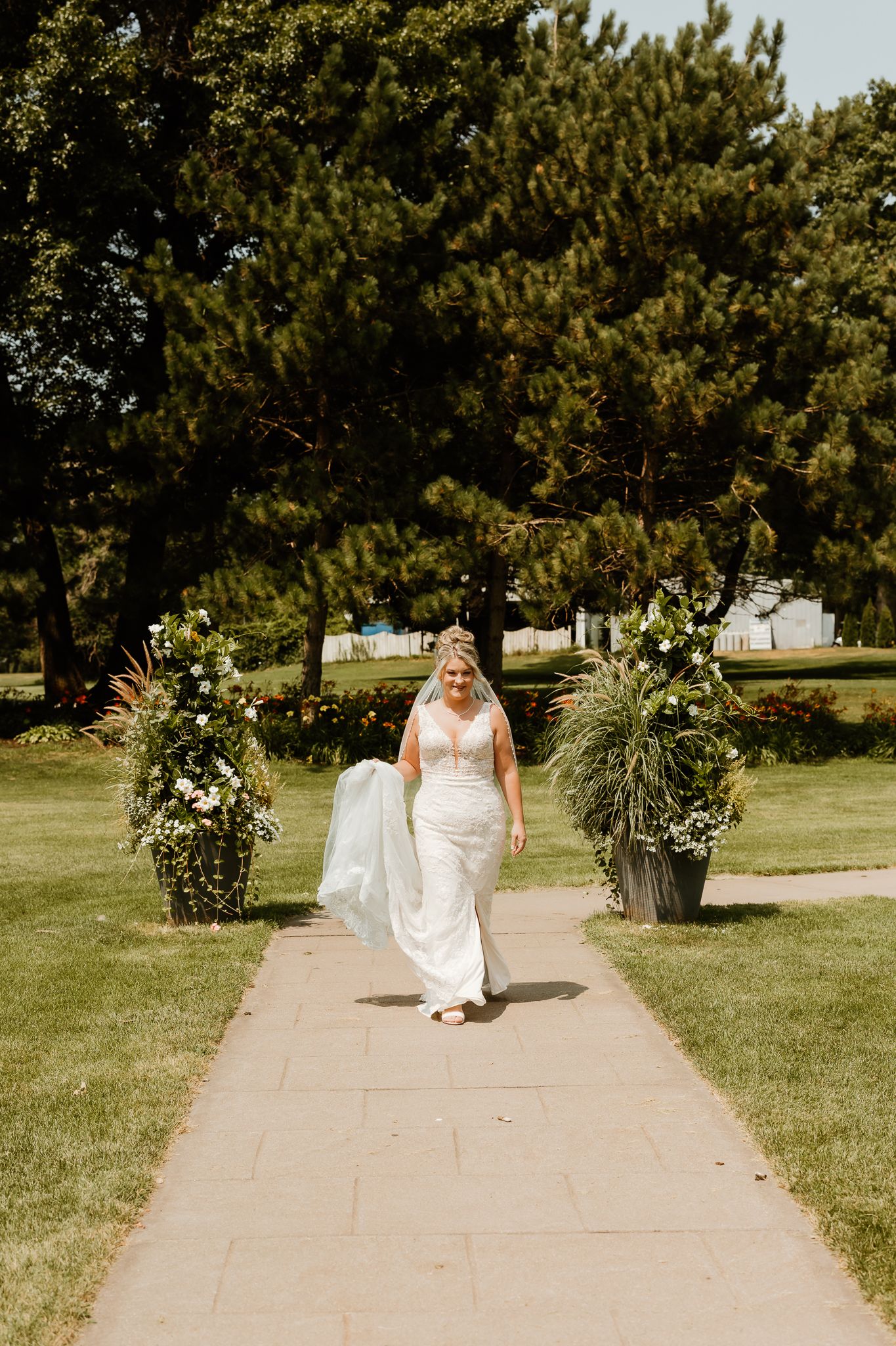 A radiant bride in a white gown walks down a garden pathway flanked by lush floral arrangements on a sunny day.
