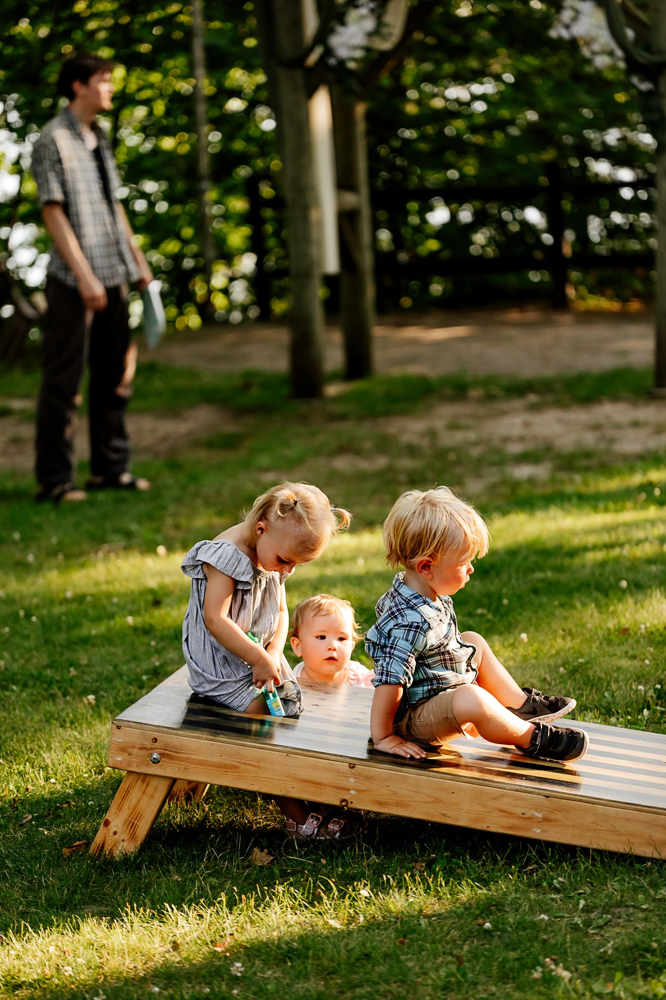 Three young children playing on a wooden cornhole board in a sunny park while an adult supervises in the background.