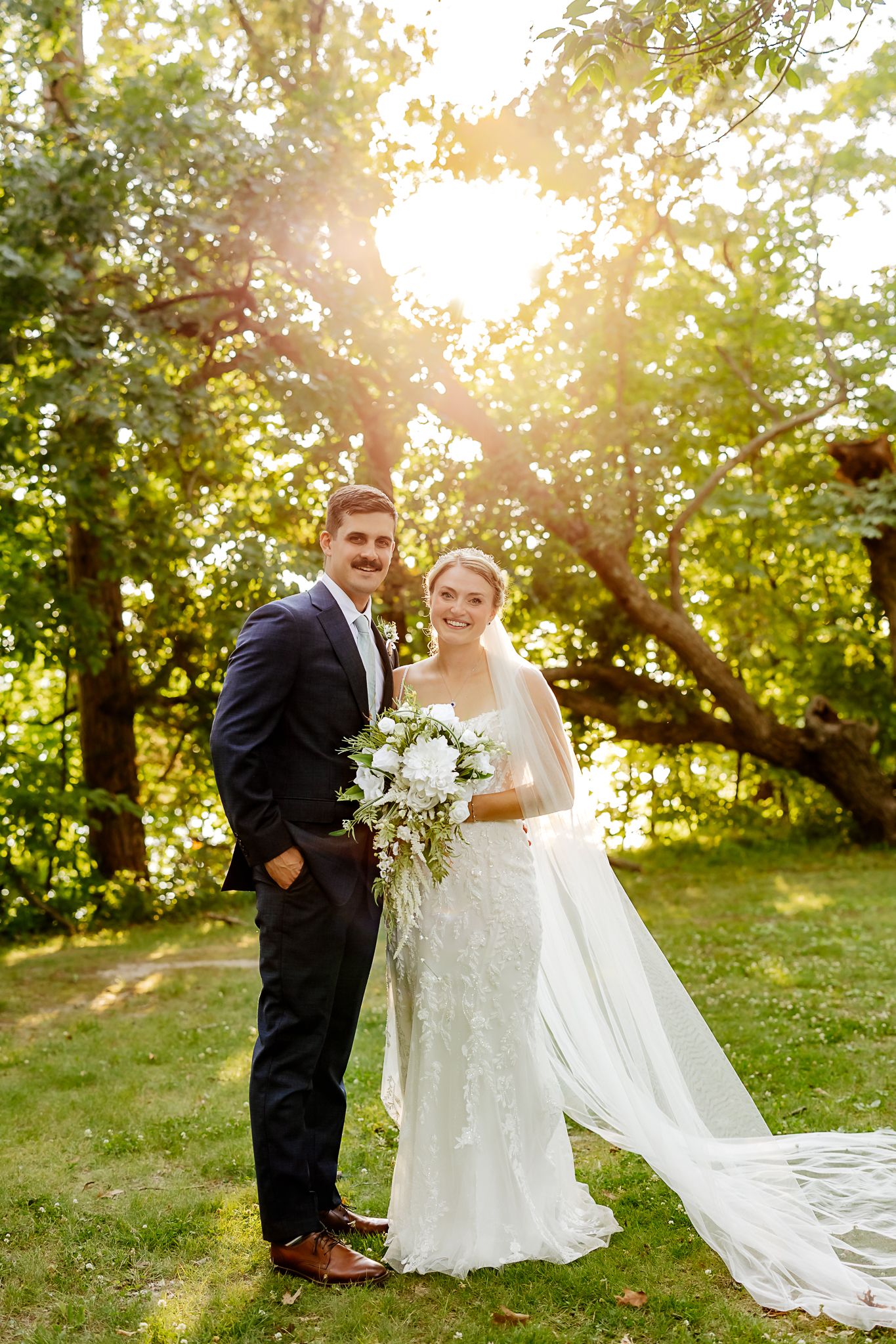 Bride and groom posing outdoors in golden sunlight, bride holding white bouquet with flowing veil, groom in navy suit.