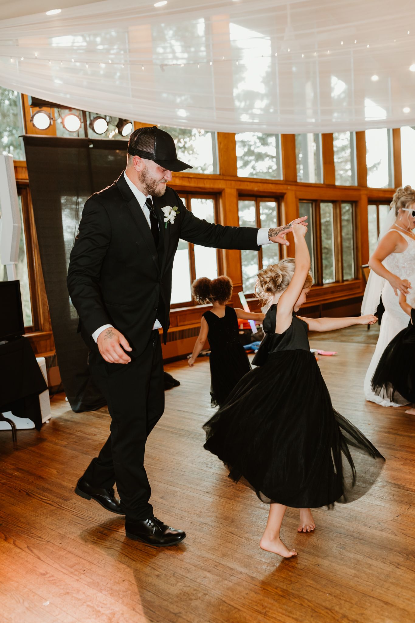 Groom in black suit and hat dancing with young flower girl in black dress at wedding reception.
