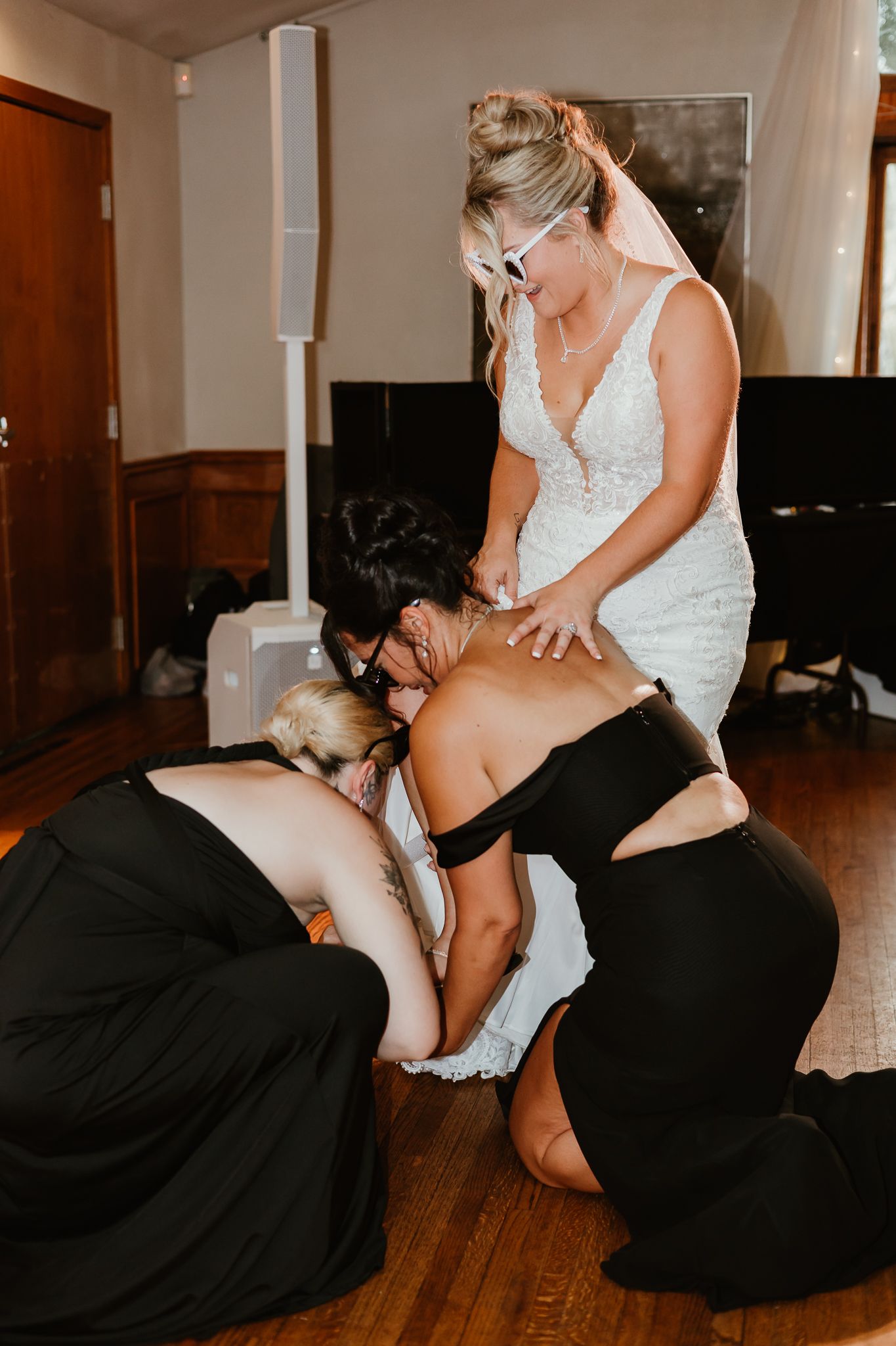 Bridesmaids in black dresses kneel to help bride in white lace gown with dress adjustments on wooden floor.