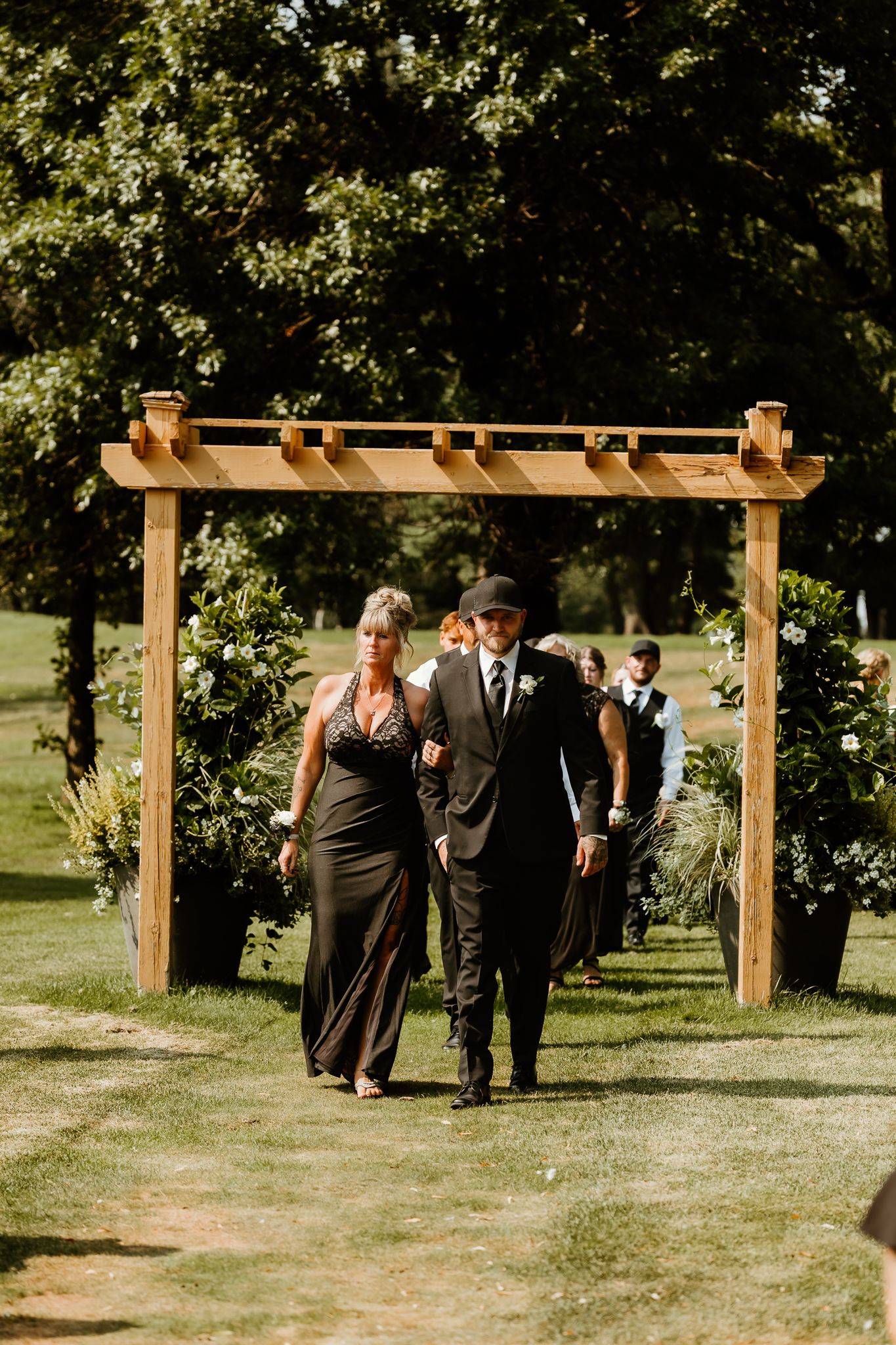 Groom in suit escorts woman in black gown down outdoor wedding aisle under wooden pergola arch.