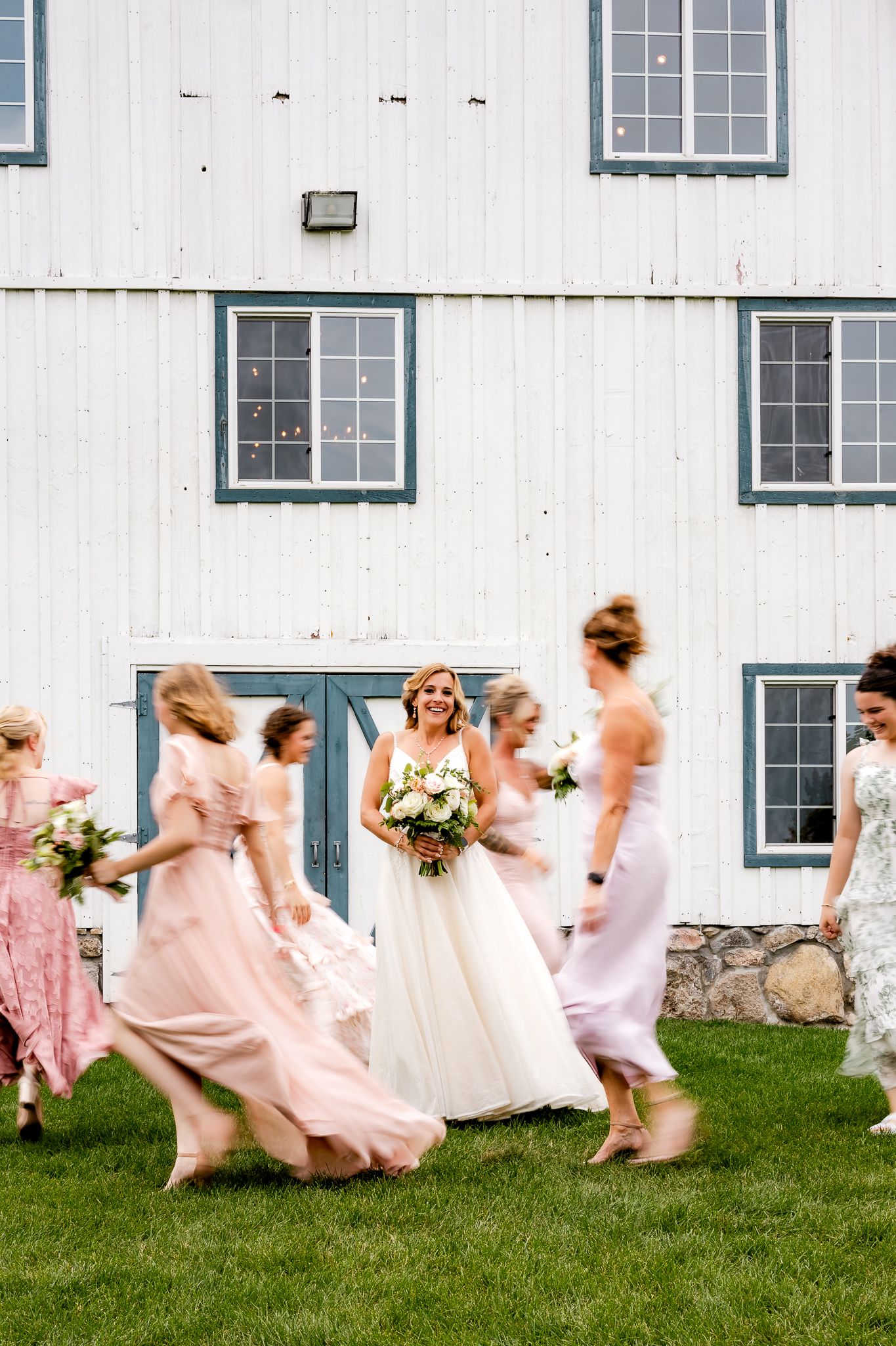 Bride in white gown walks with bridesmaids in pink dresses outside a white barn venue on a sunny day.
