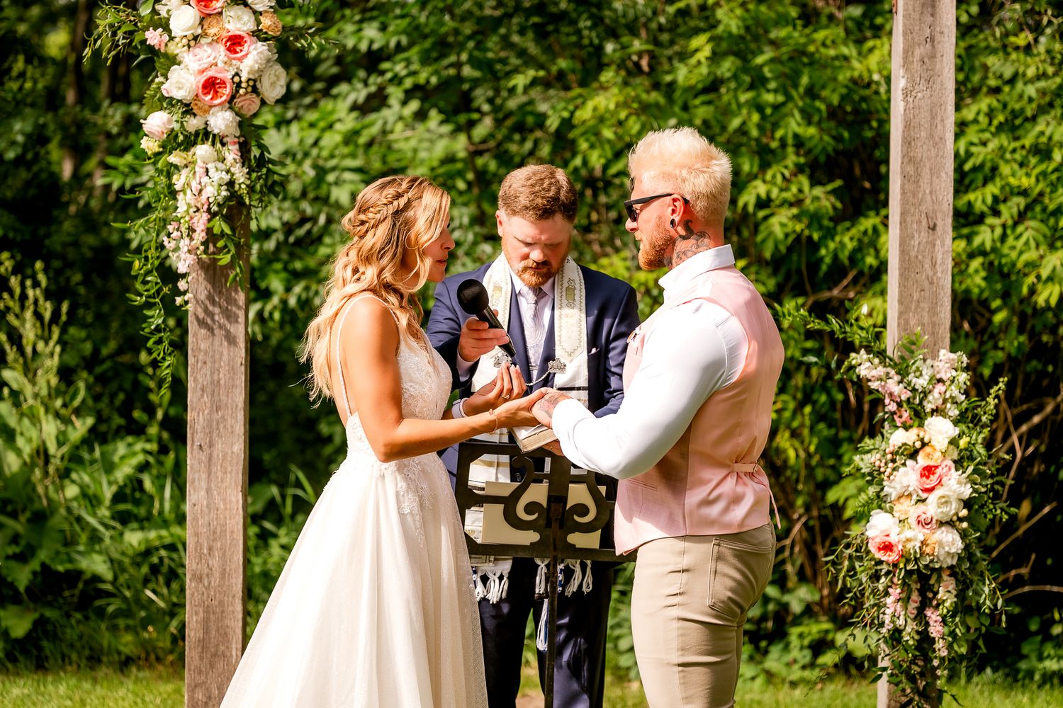 Outdoor wedding ceremony with bride and groom exchanging rings before officiant, framed by wooden arch with pink floral arrangements.