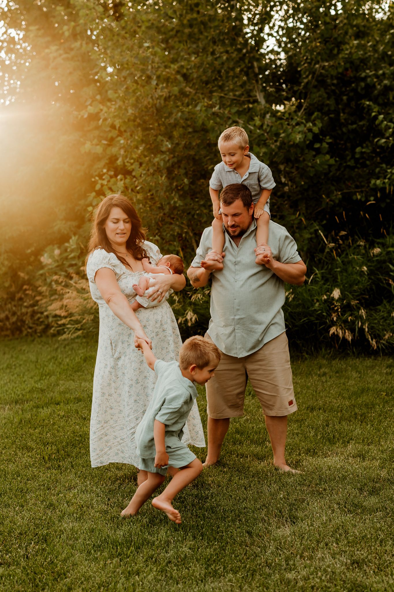 Family of five playing outdoors at golden hour, mom holding newborn, dad with toddler on shoulders, child tumbling on green grass.